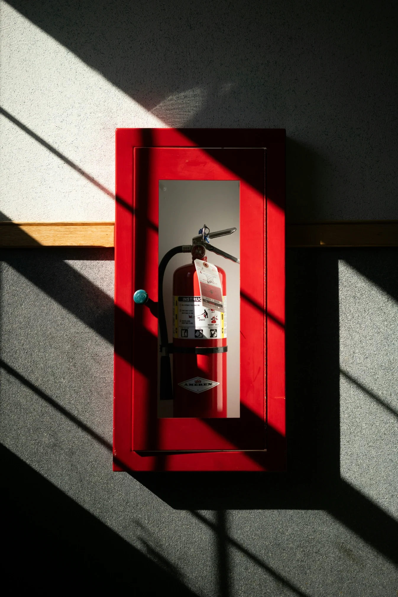 Red fire extinguisher in a red metal box with a mirror on the front, mounted on a gray wall, with sunlight casting shadows across the wall.