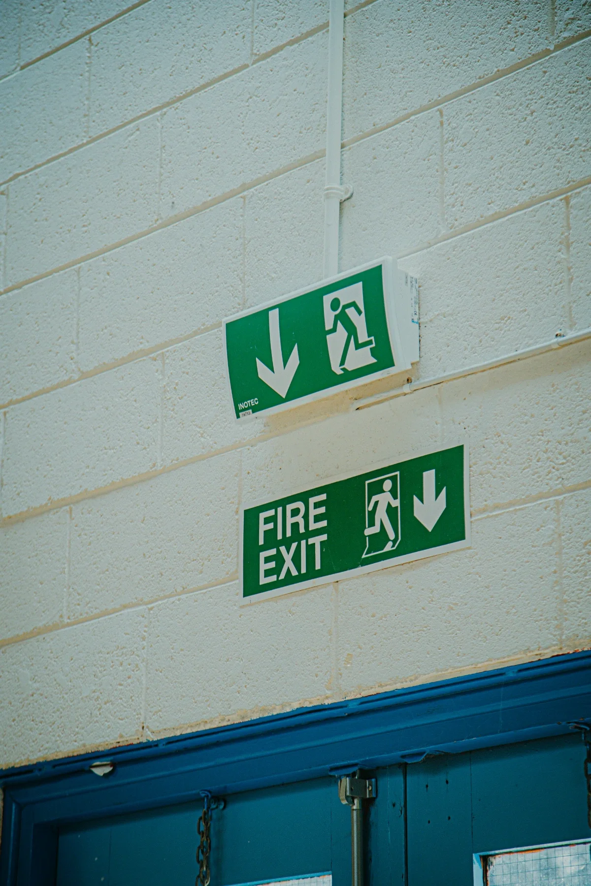 Green emergency exit signs on a beige cinder block wall, indicating the direction downward for evacuation, with icons of a person running through a door, and labeled 'FIRE EXIT'.