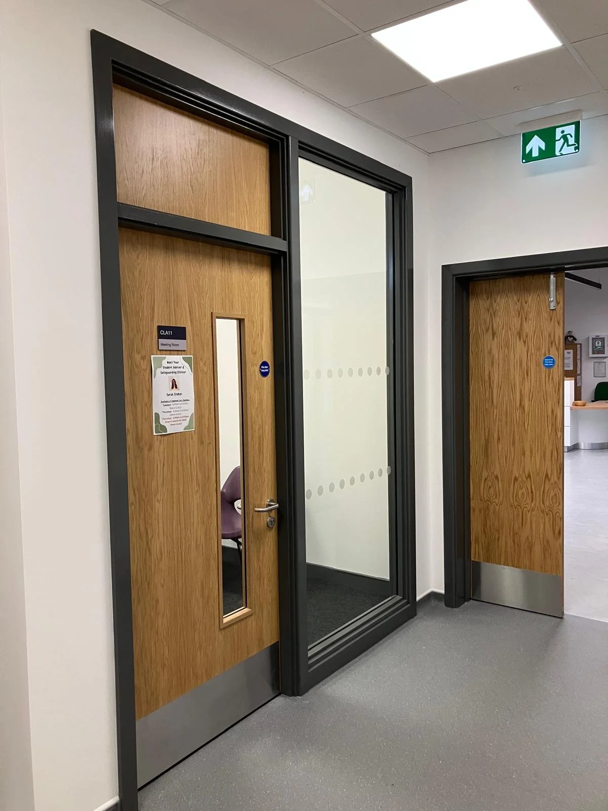 Wooden doors in a hallway, one door has a glass window and a sign, and there is a small poster on it. The other door is partly open, revealing an office area. An illuminated green emergency exit sign is above, indicating the way out.