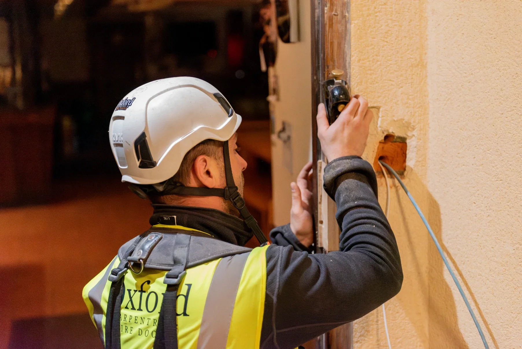 A construction worker wearing a helmet and safety vest installing or repairing electrical wiring in a wall.