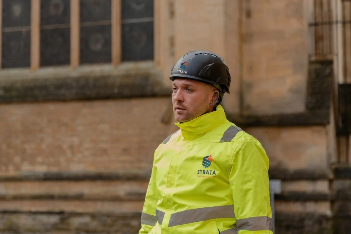 A firefighter wearing a black helmet and a bright yellow jacket with reflective stripes, standing outdoors in front of a brick building.