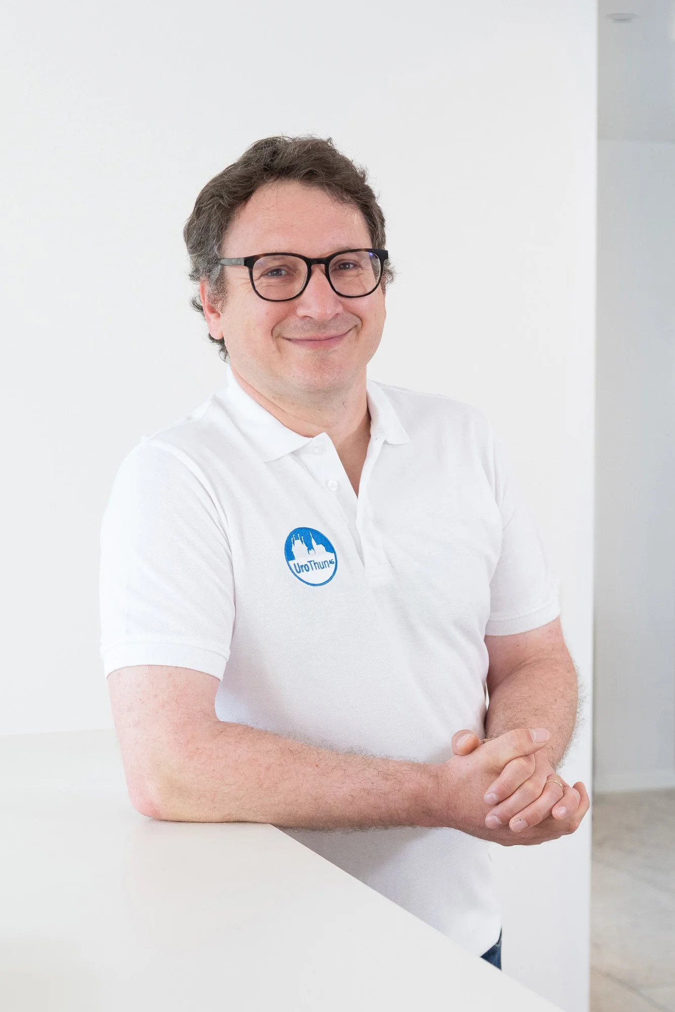 Mann in weißem Polo mit Brille und Lächeln, steht vor einem weißen Hintergrund, Arme verschränkt auf einem weißen Tisch, Logo auf der Brust.