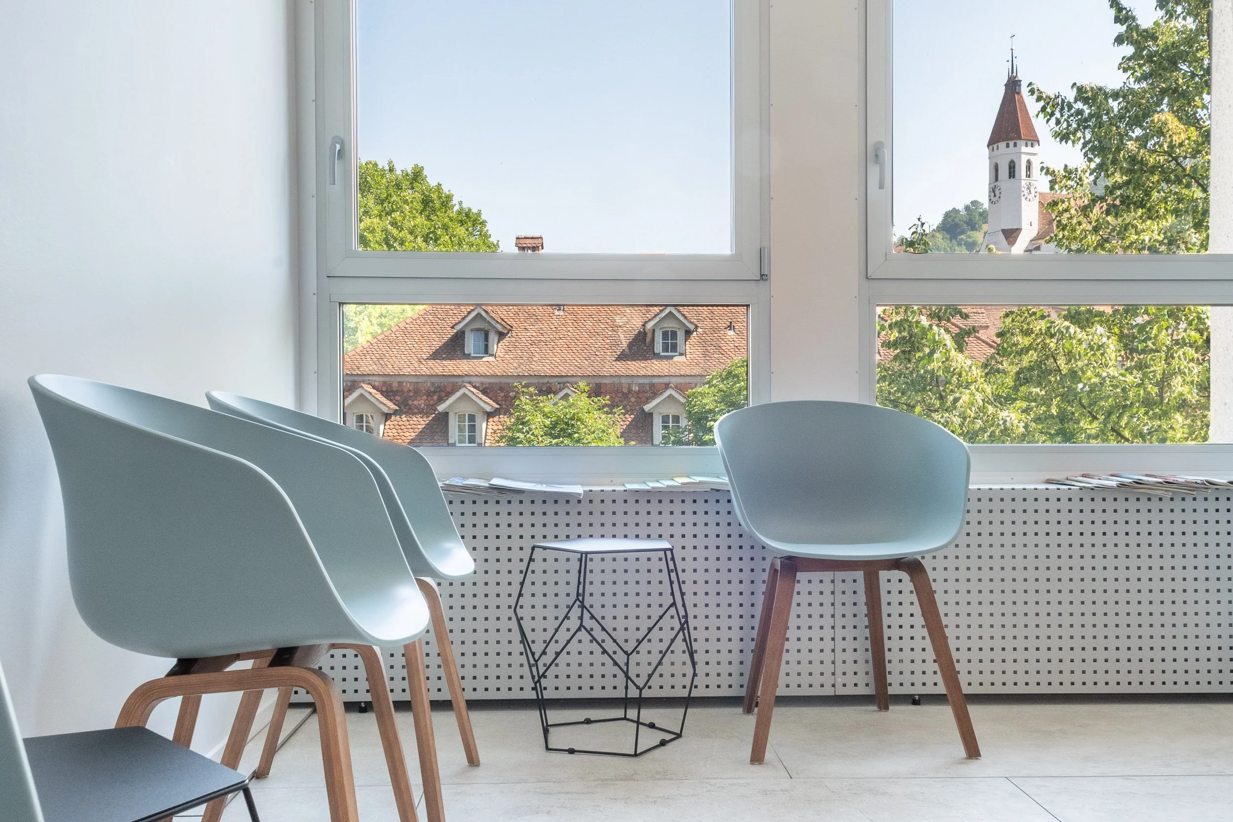 Innenraum eines modernen Warteraums mit weißen Stühlen, einem schwarzen Beistelltisch, großen Fenstern und Blick auf ein Kirchtürmchen und Dächer im Freien.