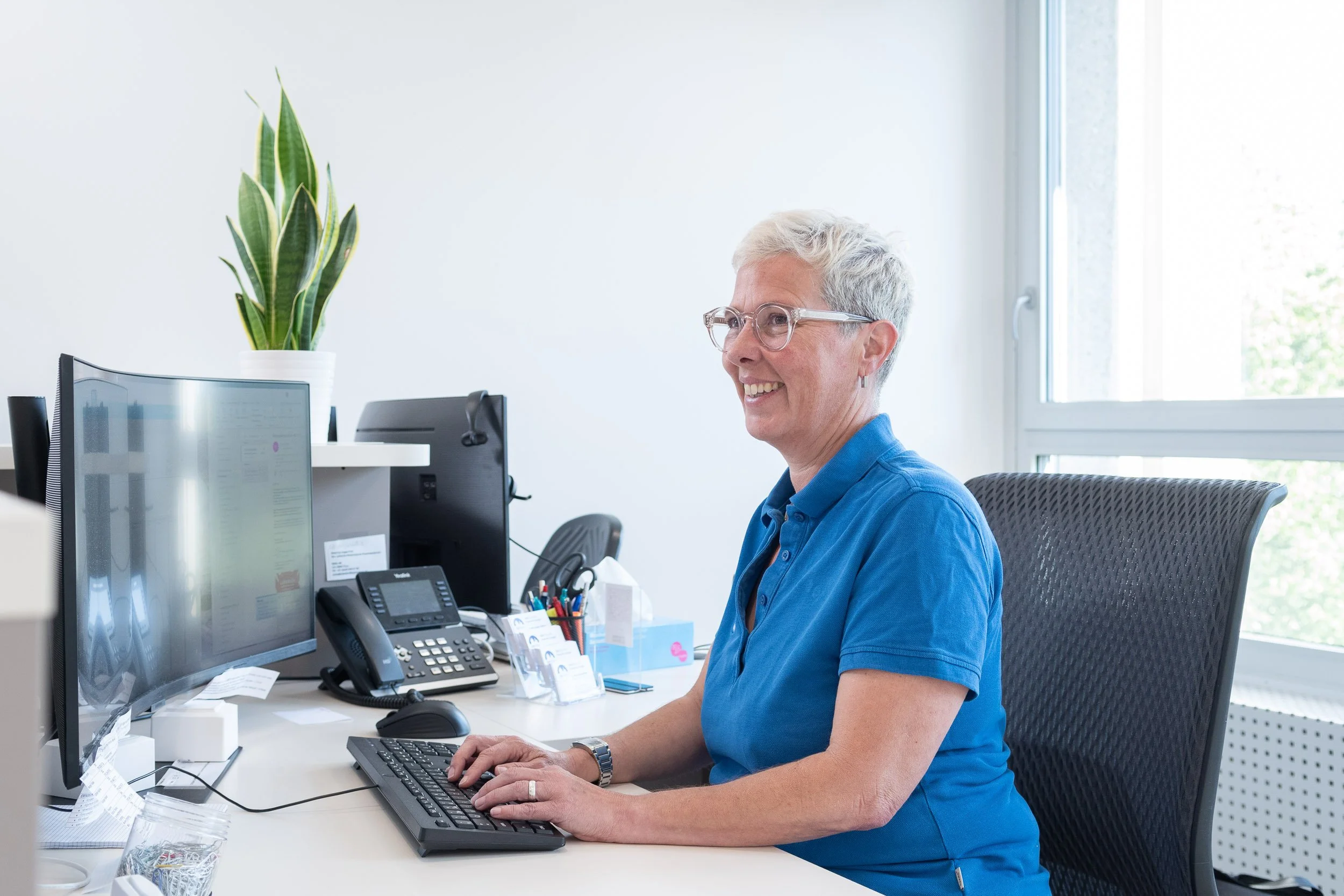 Eine Frau mit kurz grauem Haar, Brille und blauer Bluse sitzt an einem Schreibtisch im Büro, arbeitet am Computer und lächelt.