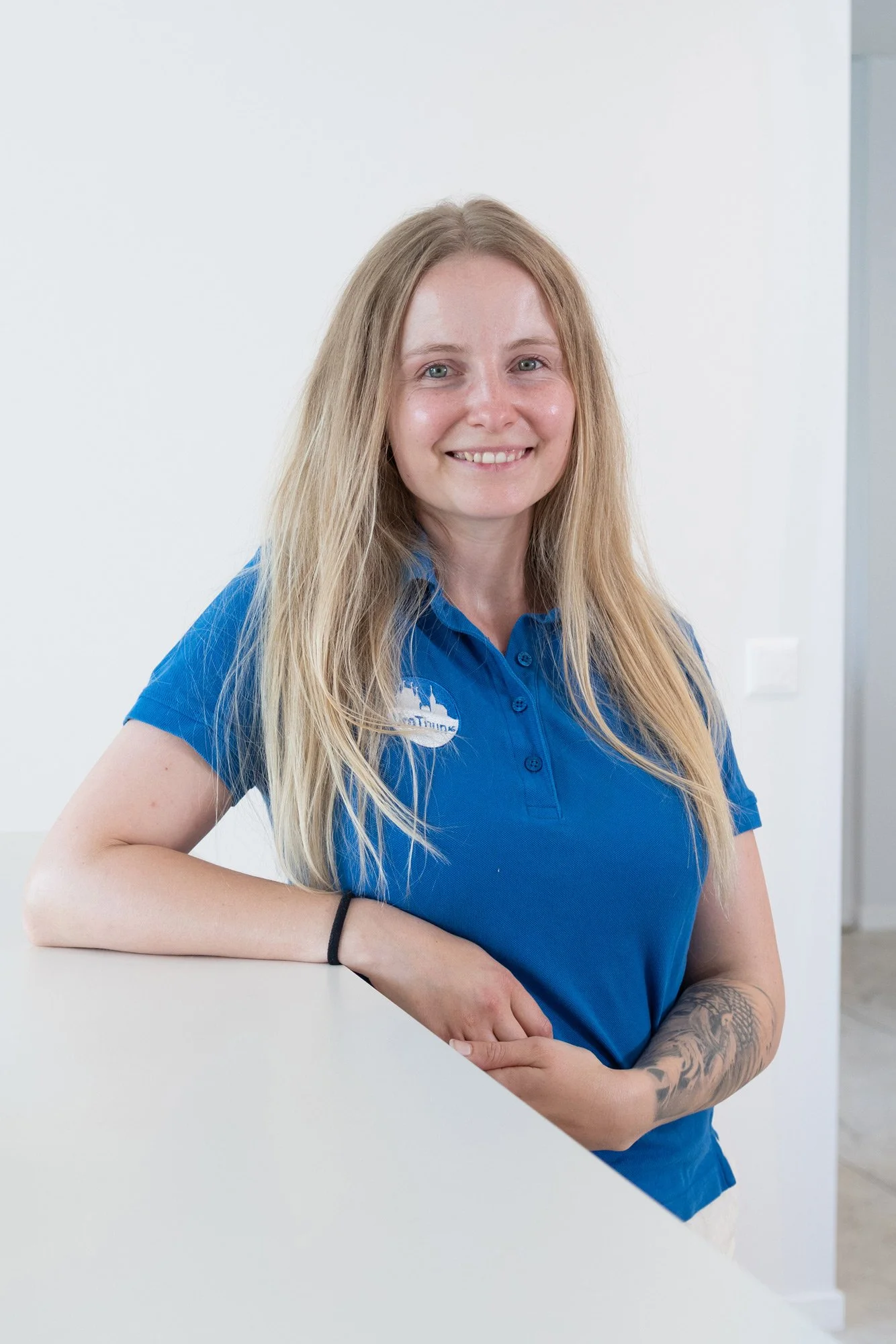 Junge Frau mit langem blondem Haar, blauer Polo, Arm mit Tattoo, steht an einem weißen Tresen, lächelt und schaut in die Kamera.