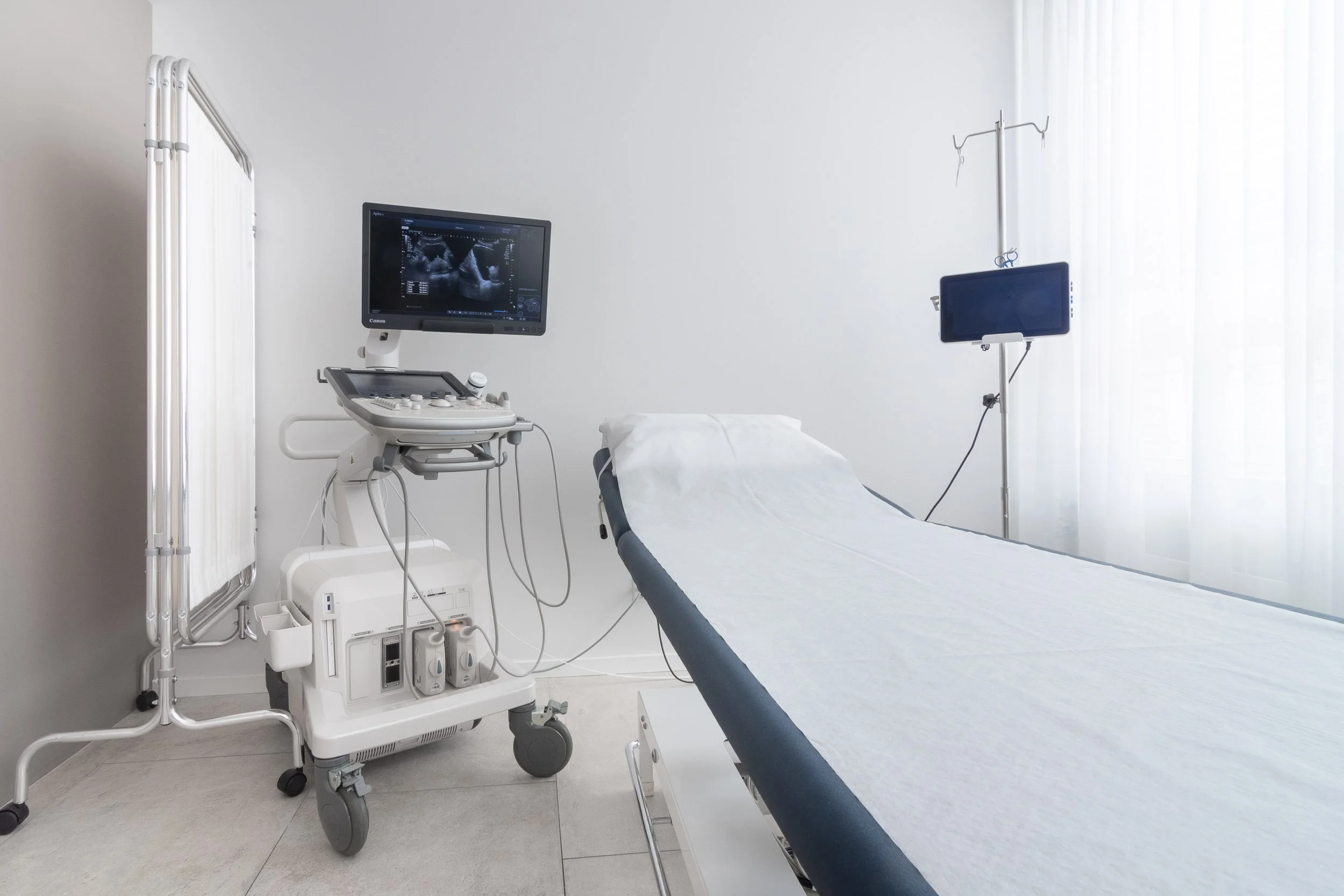 Ein dunkelblauer Untersuchungstisch mit weißen Bettlaken in einem weißen, leeren Krankenhauszimmer neben einem Ultraschallgerät und einem Monitor, der eine Ultraschallaufnahme zeigt.