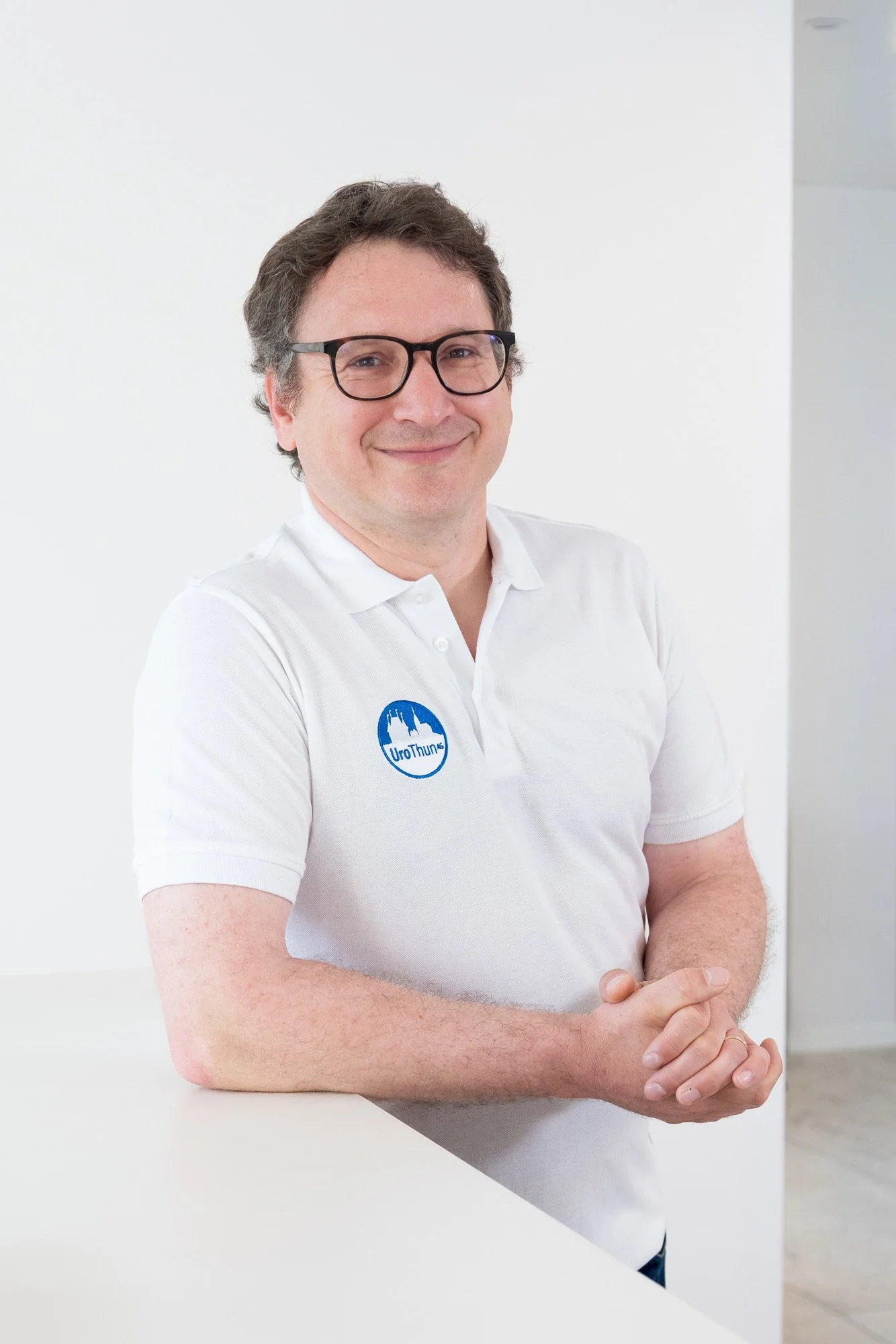 Mann mit Brille trägt ein weißes Polo-Shirt mit einem Logo, lächelt, steht vor einer weißen Wand, die sein Oberkörper einschließt.