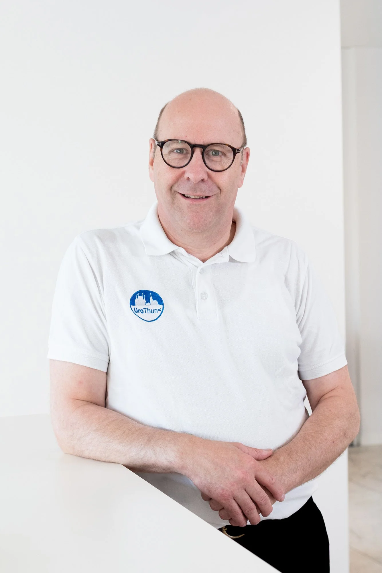 Mann trägt ein weißes Polo Shirt mit einem Logo, steht vor weißer Wand und lächelt.