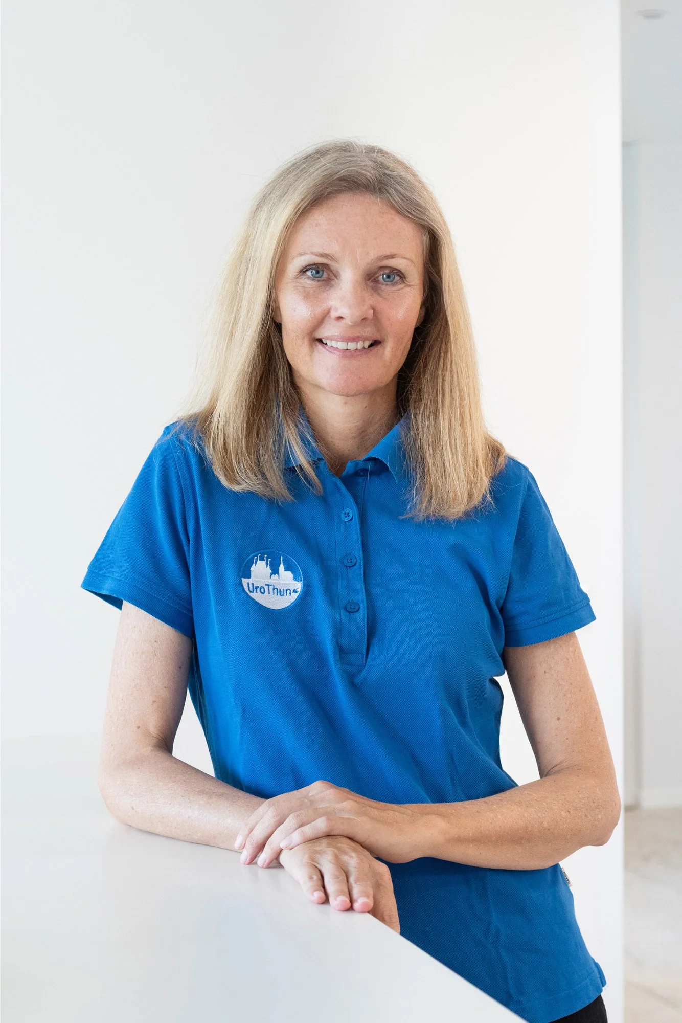 Frau mit blauer Polo-Hemd, sitzt an einem weißen Tisch, lächelt, hat lange blonde Haare und blaue Augen.