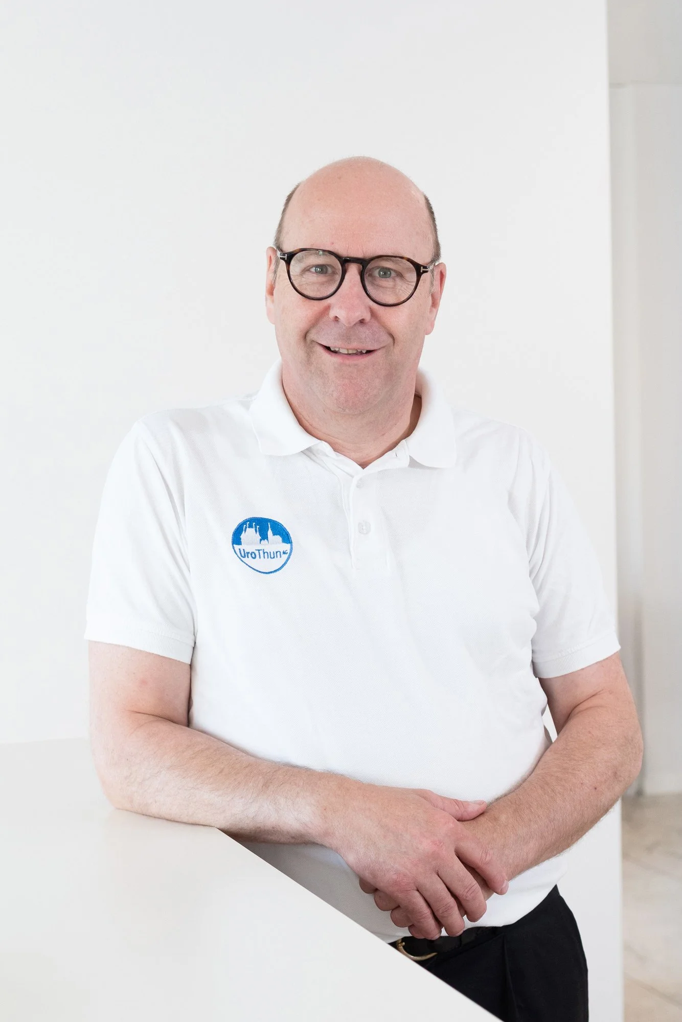 Ein Mann mit Brille und weißem Polo, der vor einem weißen Hintergrund steht und in die Kamera lächelt.