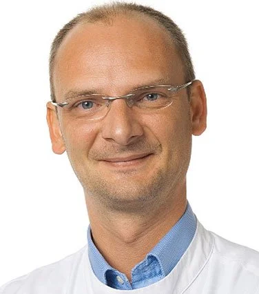 Porträt eines Mannes mit Brille, kurz geschnittenen Haaren und einem weißen Arztkittel.