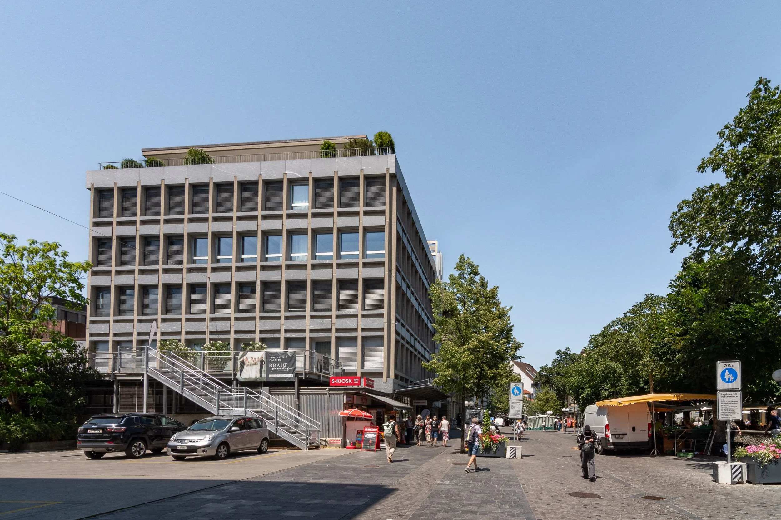 Ein mehrstöckiges Gebäude mit modernen Fenstern und einer Dachterrasse, umgeben von Bäumen, auf einem belebten öffentlichen Platz mit Menschen und Fahrzeugen, Laden und Informationsschildern.