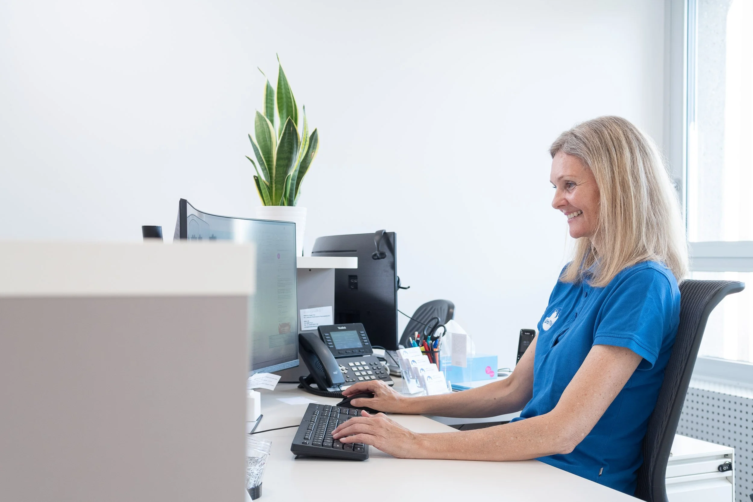 Eine lächelnde Frau sitzt an einem Schreibtisch im Büro, arbeitet am Computer, umgeben von Bürobedarf, Telefon und einer Pflanze auf einem Regal im Hintergrund.