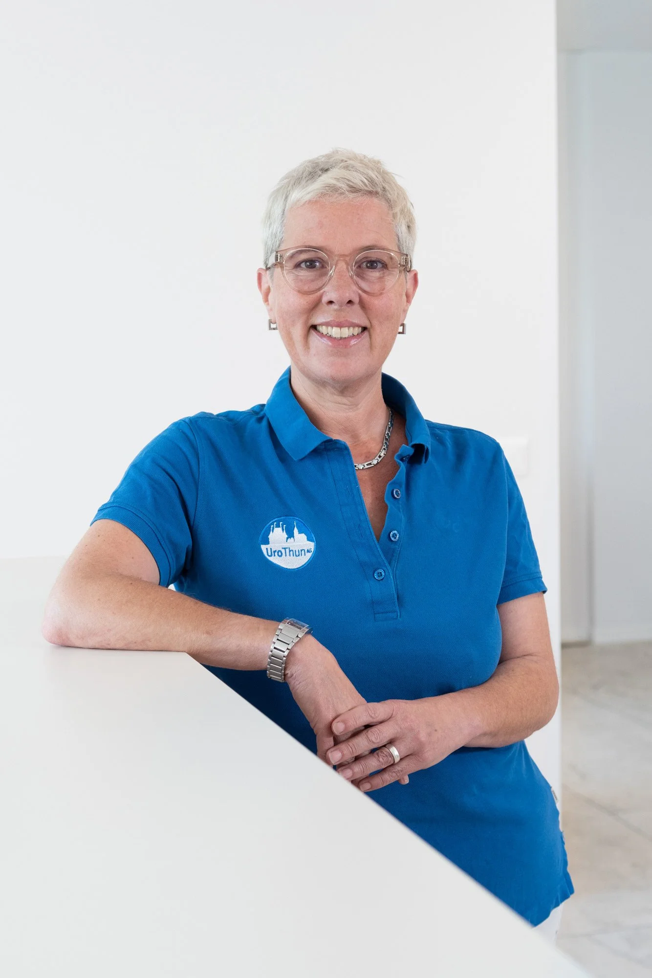 Lächelnde Frau mit kurzen hellen Haaren, Brille, blauer Polo mit Logo, steht in hellen Raum, Arm auf Tisch, Blick in Kamera.