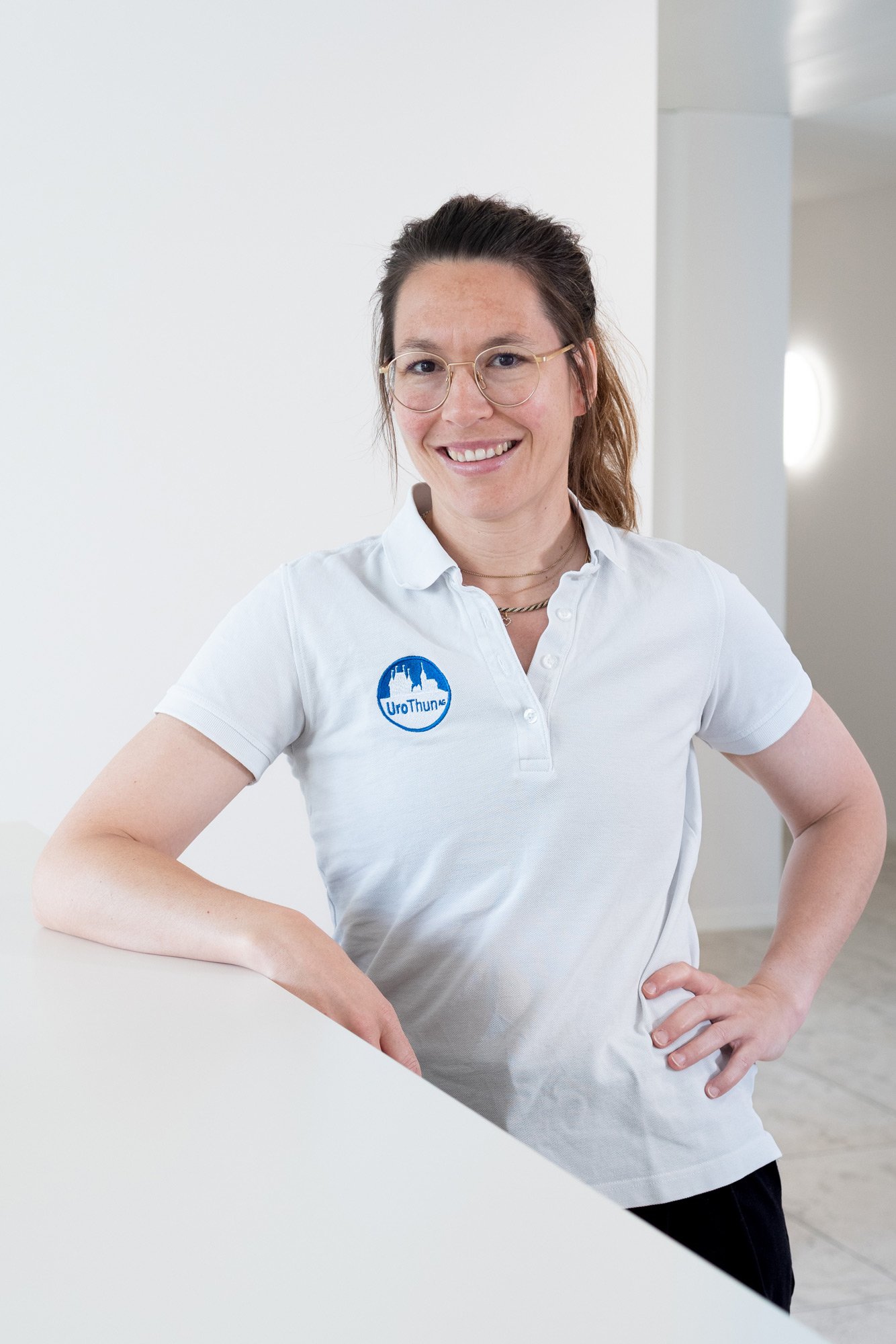 Junge Frau mit Brille und weißen Polo-Shirt, die vor einer weißen Wand steht, lächelt und die Hände auf die Hüften legt.
