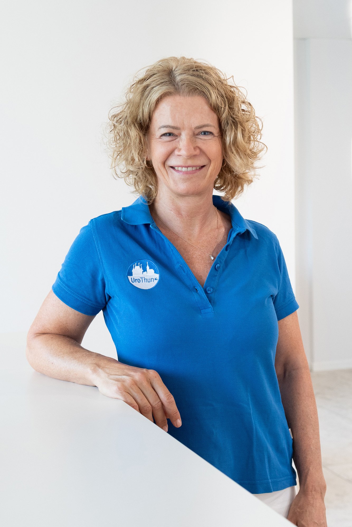 Eine lächelnde Frau in blauer Polo-Hemd mit einem UroThun Logo, die an einer weißen Wand lehnt.