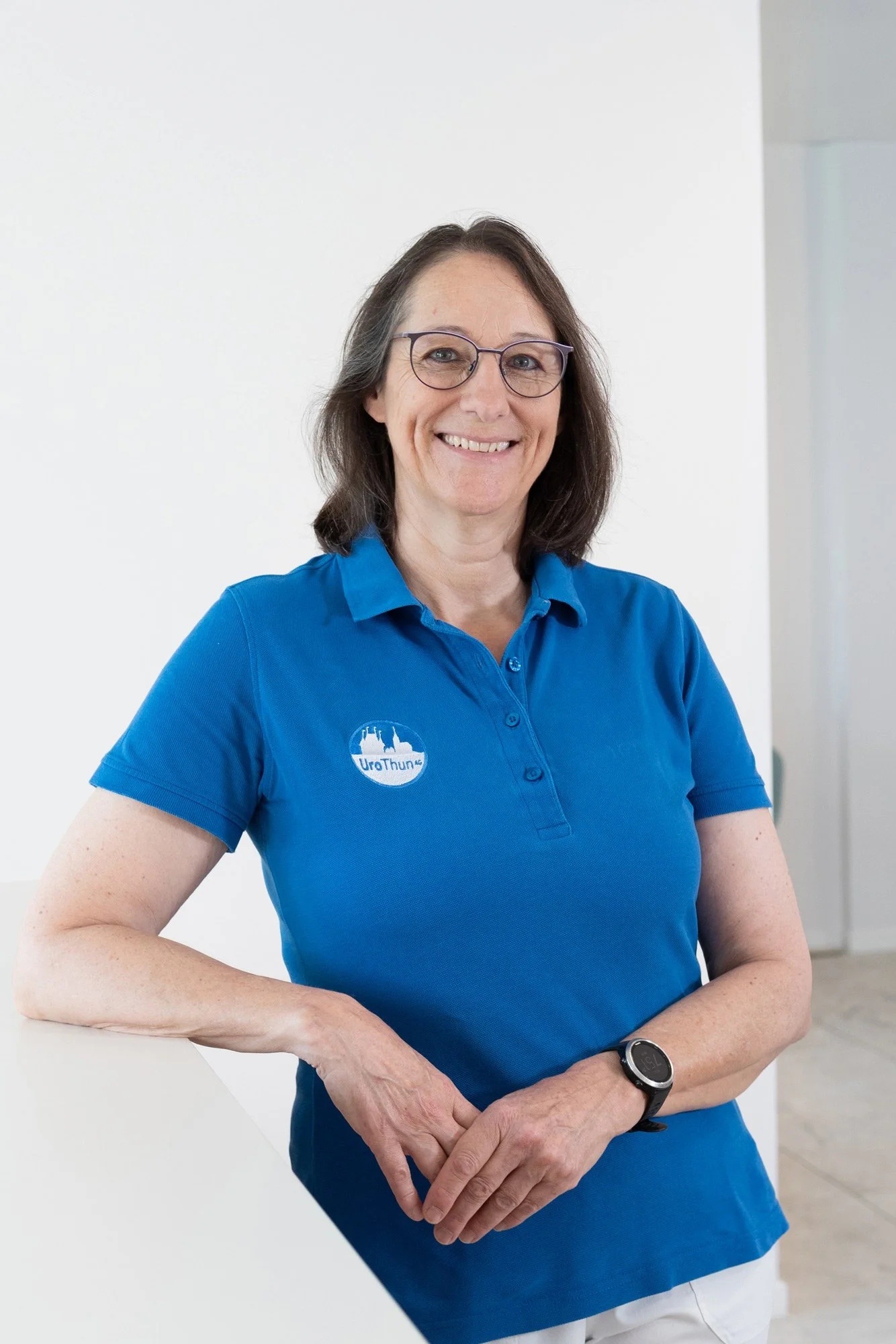 Frau mit Brille und blauer Polo-Shirt steht vor weißer Wand, lächelt, trägt eine Armbanduhr und hat eine Logo auf dem Polo-Shirt.