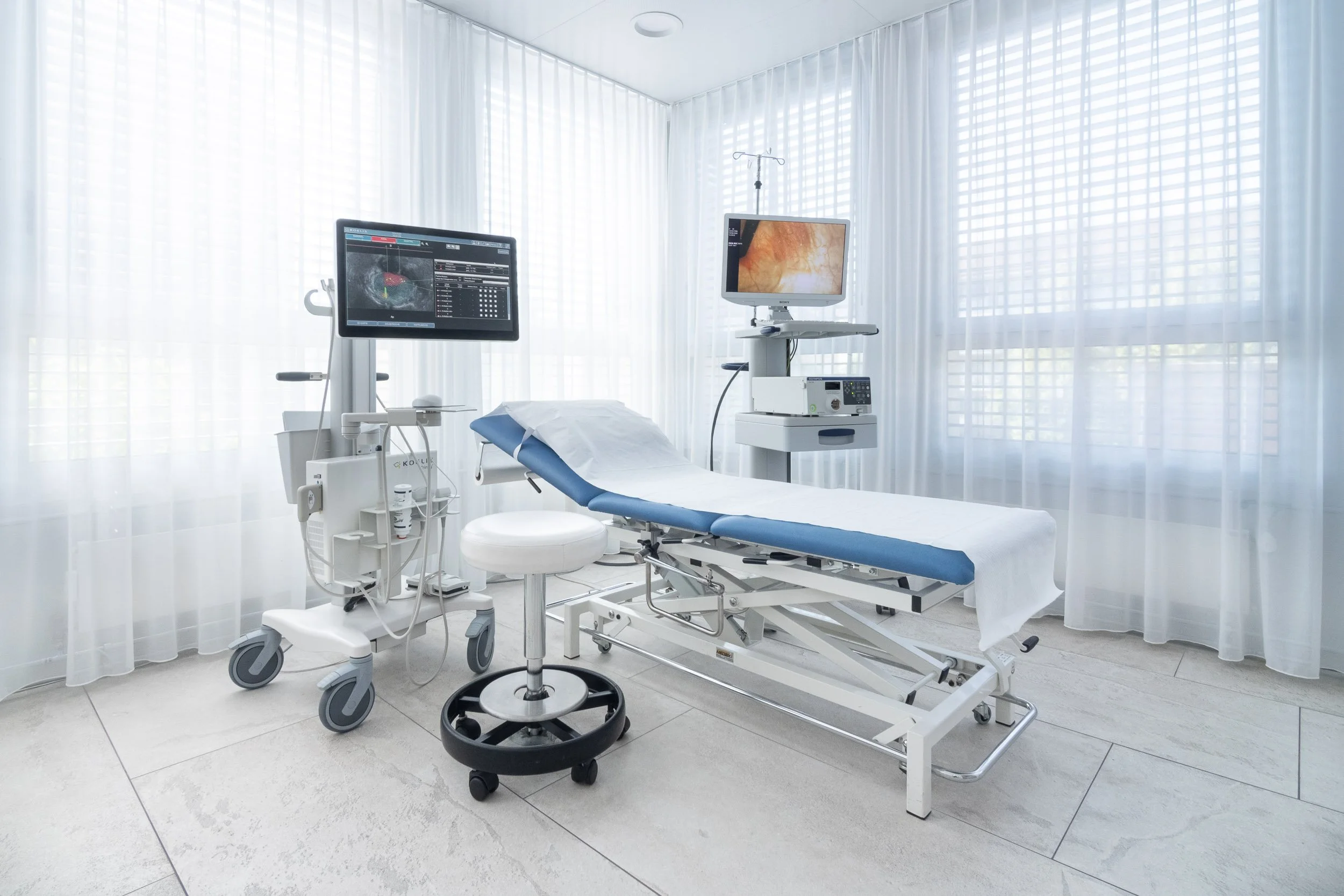 Ein Krankenhauszimmer mit einem Untersuchungsliege, Monitoren und medizinischer Ausrüstung, das helle, lichtdurchflutete Zimmer hat Fenstervorhänge.