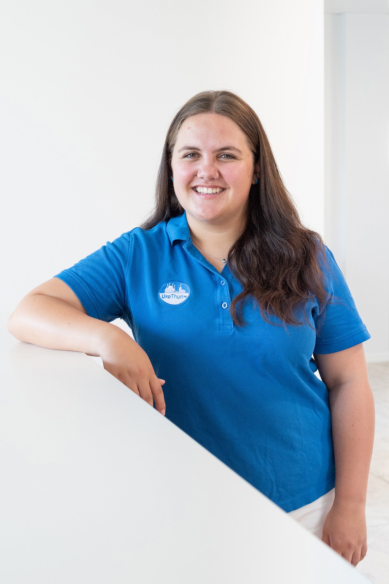 Lächelnde Frau trägt ein blaues Poloshirt mit einem Logo, steht an einem weißen Tisch und lächelt.