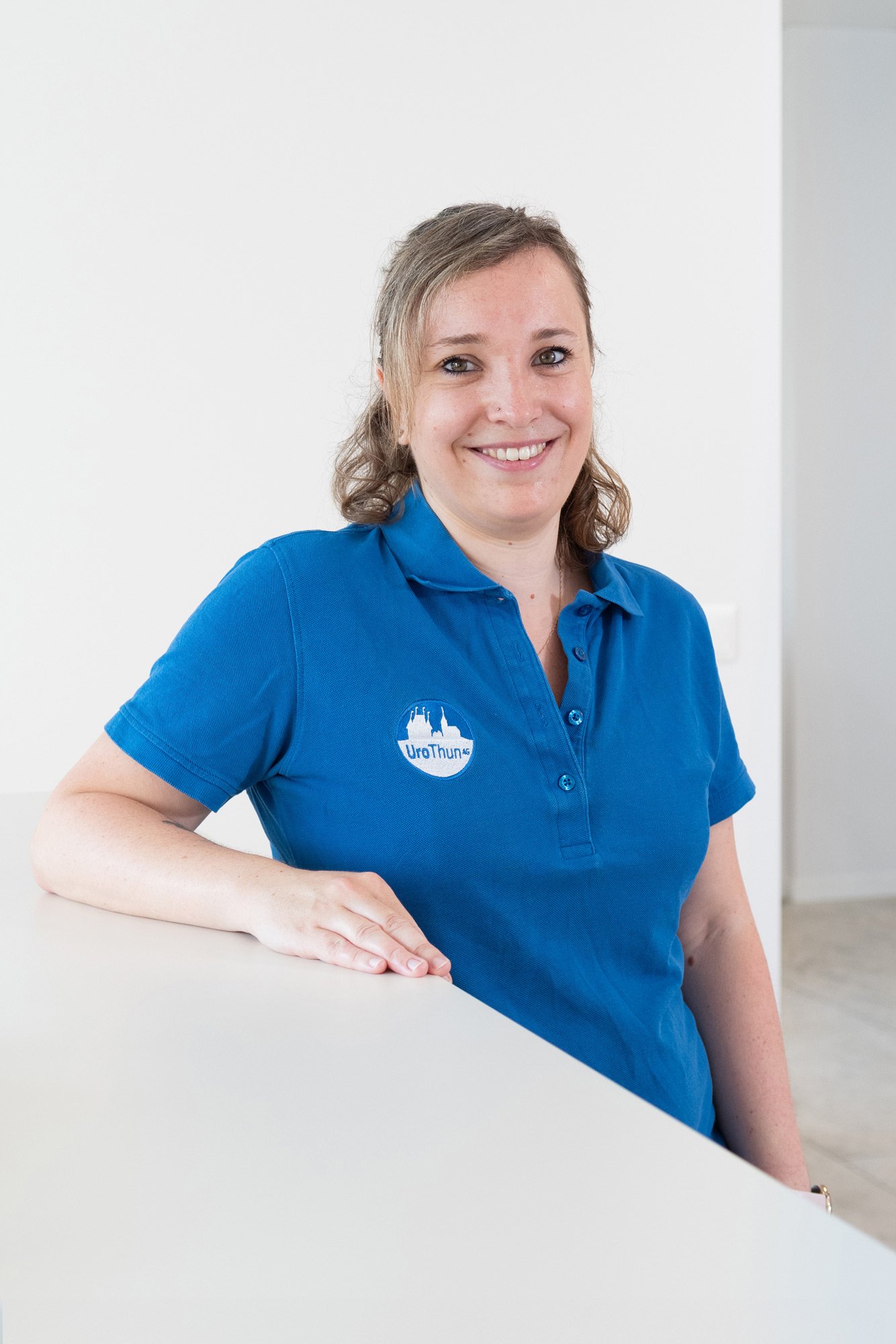 Lächelnde Frau in blauer Polo mit Logo, stehend in hellem Raum, die Hand auf einem weißen Tisch, freundlich und professionell.