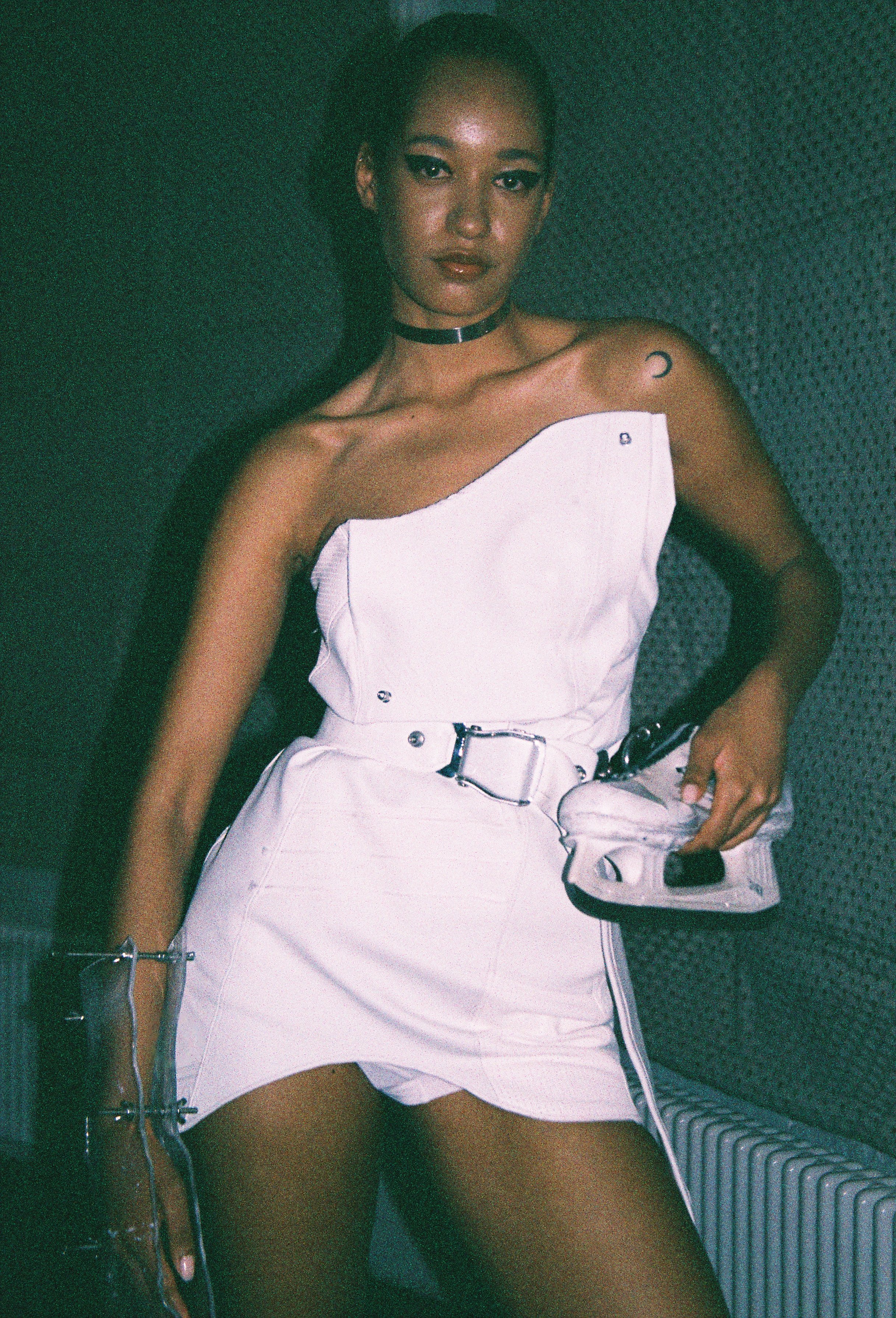 A woman lying on a textured surface, wearing a white strapless dress, choker necklace, and holding a small audio device with headphones.