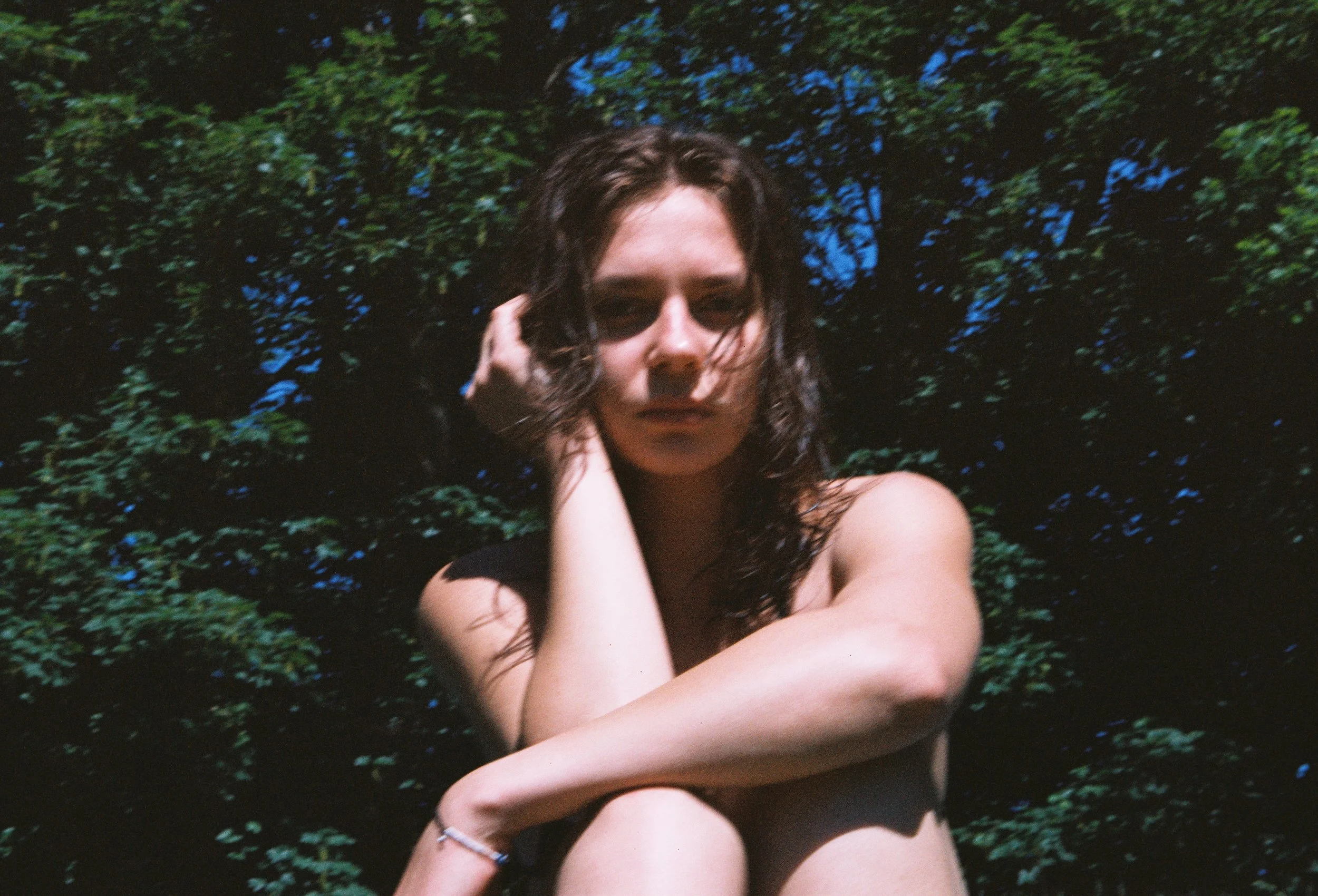 A woman with curly hair posing outdoors in front of greenery, with her hand touching her face.