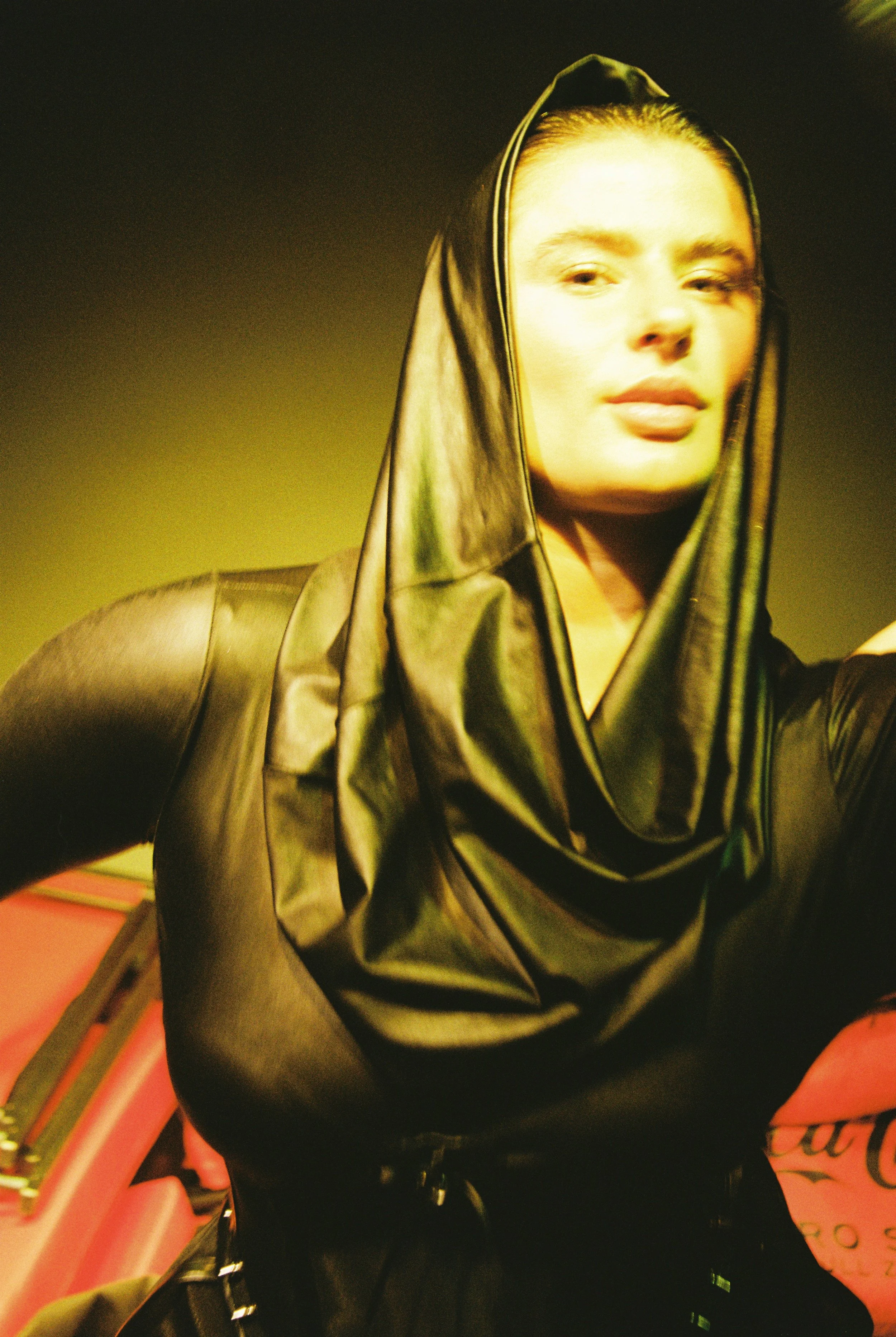 A person in a black leather hooded jacket with the hood up, sitting on a pink bench with a pink sign nearby, in an indoor setting.
