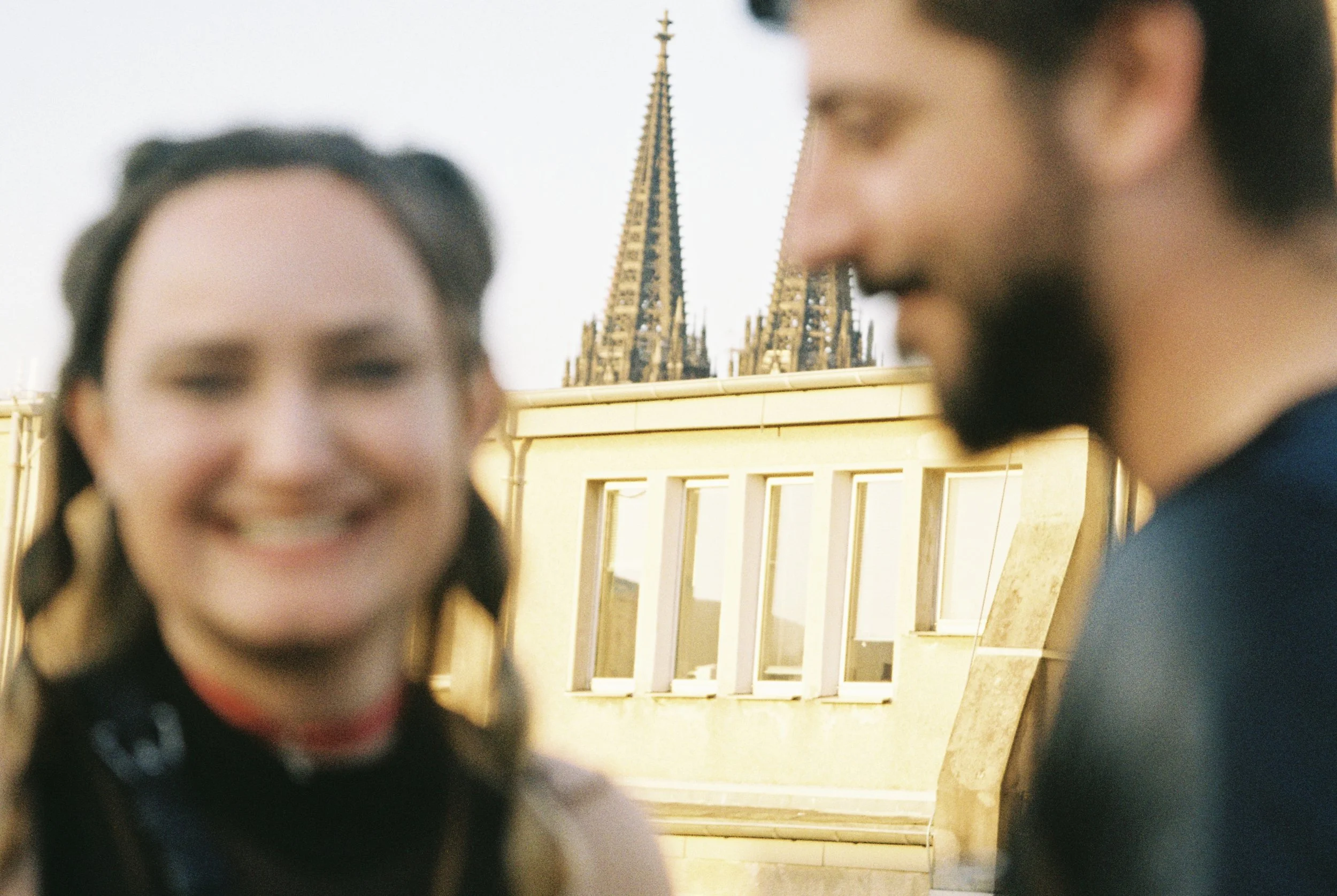 Close-up of two smiling people, a woman with dark hair in the foreground and a man with a beard and dark hair in the background, with a cathedral spire visible behind them.