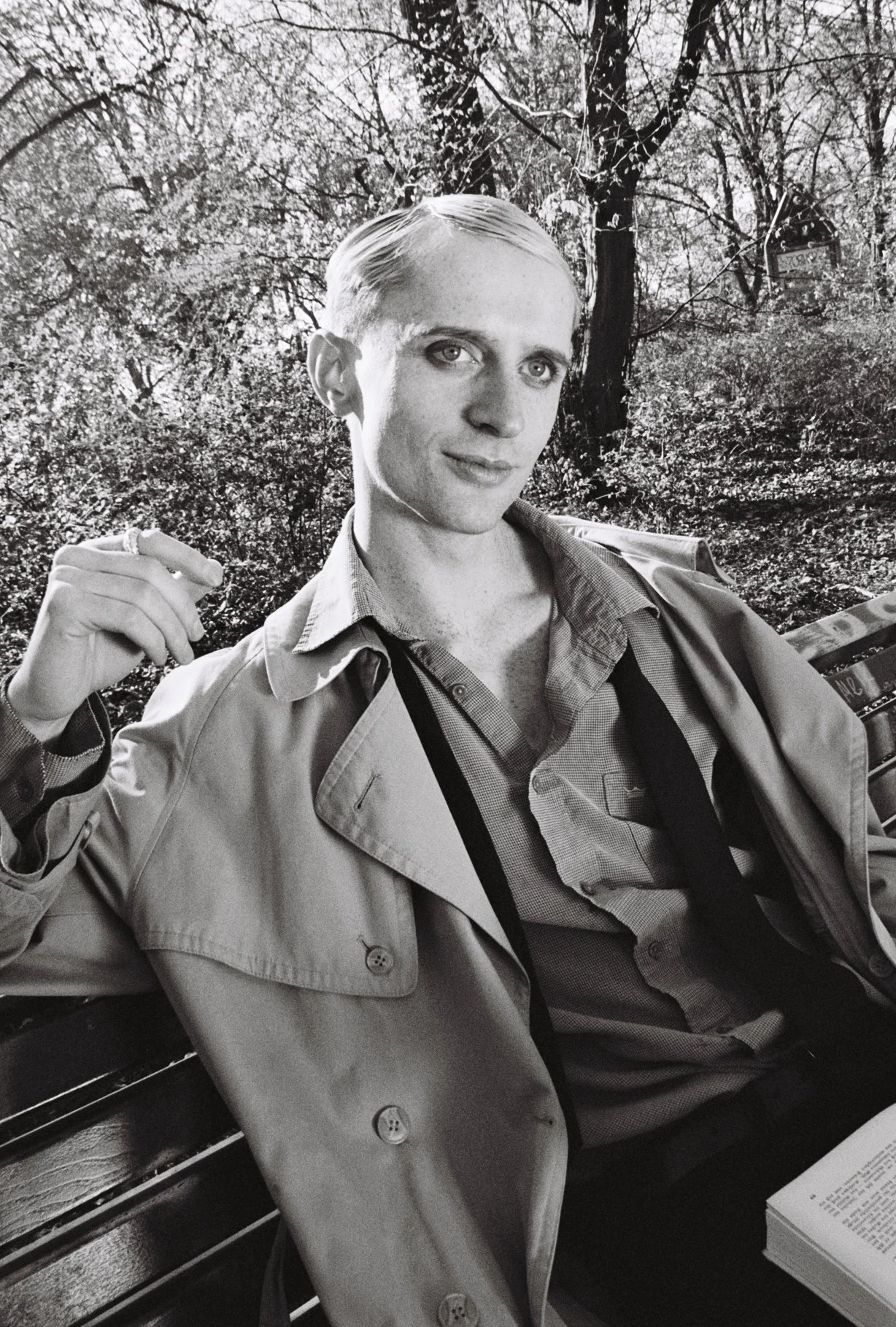 A young man with light-colored hair and fair skin sitting on a park bench surrounded by trees. He is wearing a light-colored trench coat over a shirt and has a camera strap around his neck. He is holding a cigarette in his right hand, with a slight s