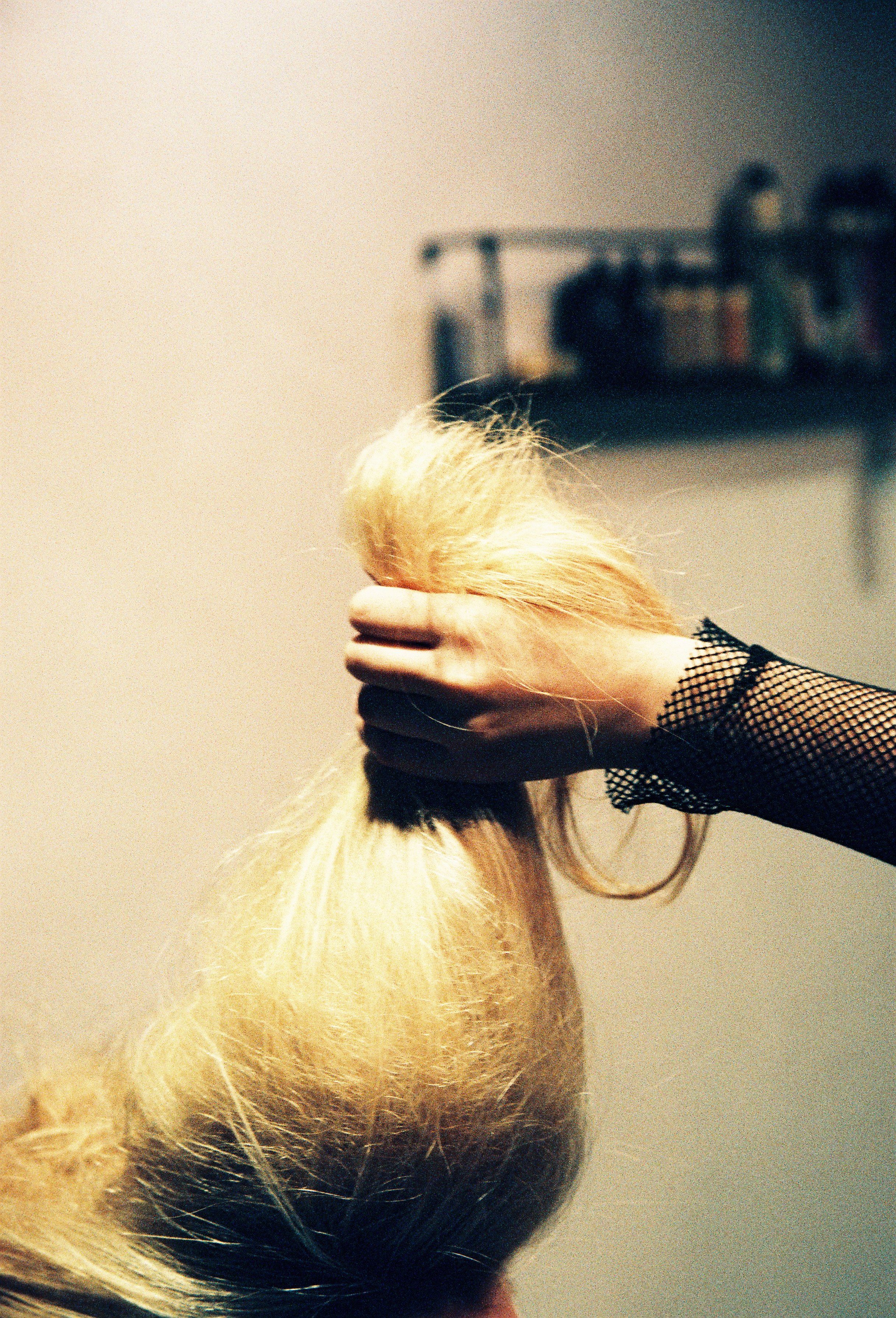 Close-up of a person holding a small blonde dog with flowing hair, wearing black mesh sleeves, against a plain tan background.