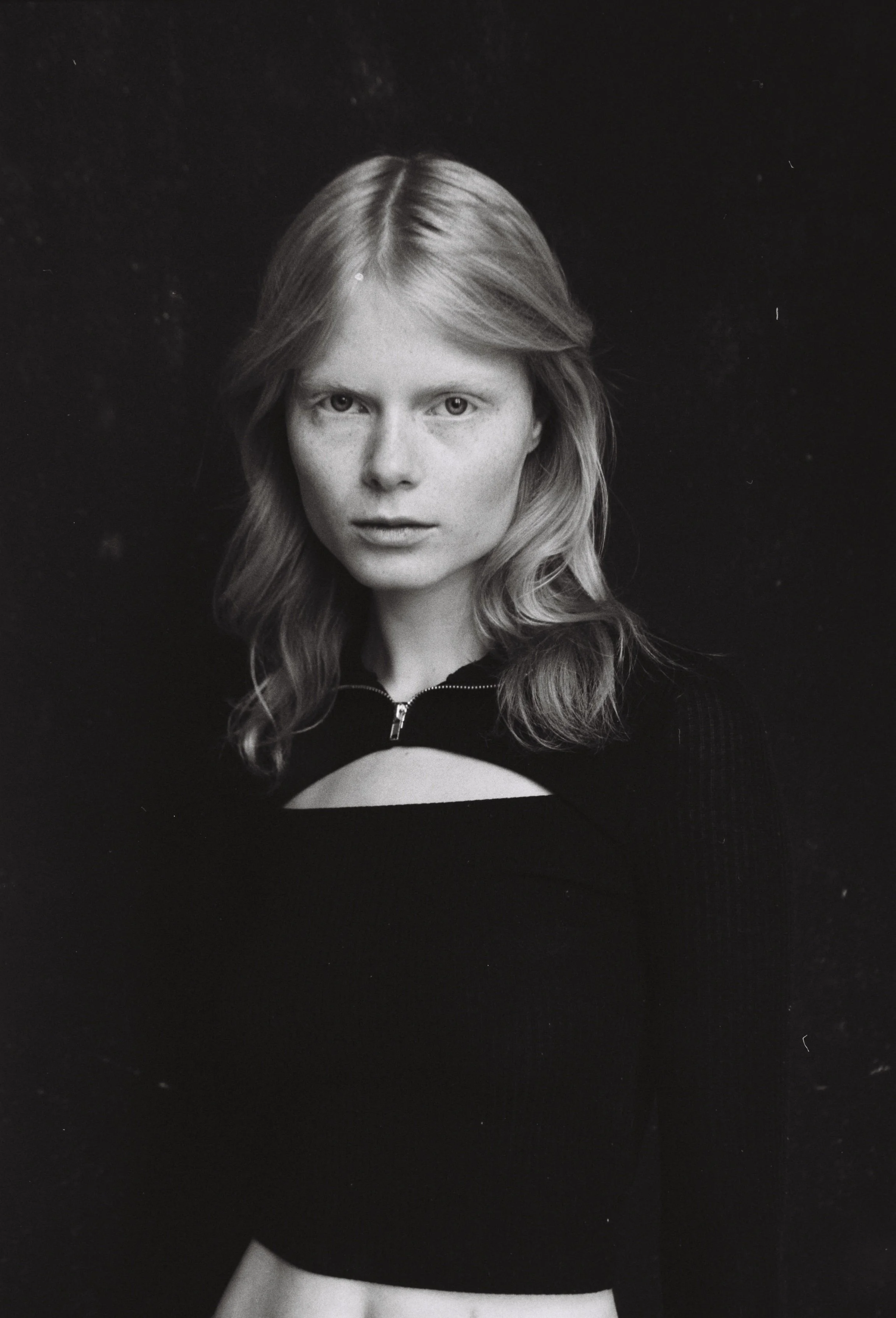 Black and white portrait of a young woman with wavy hair, wearing a zip-up top with a cut-out in the chest area, against a dark background.