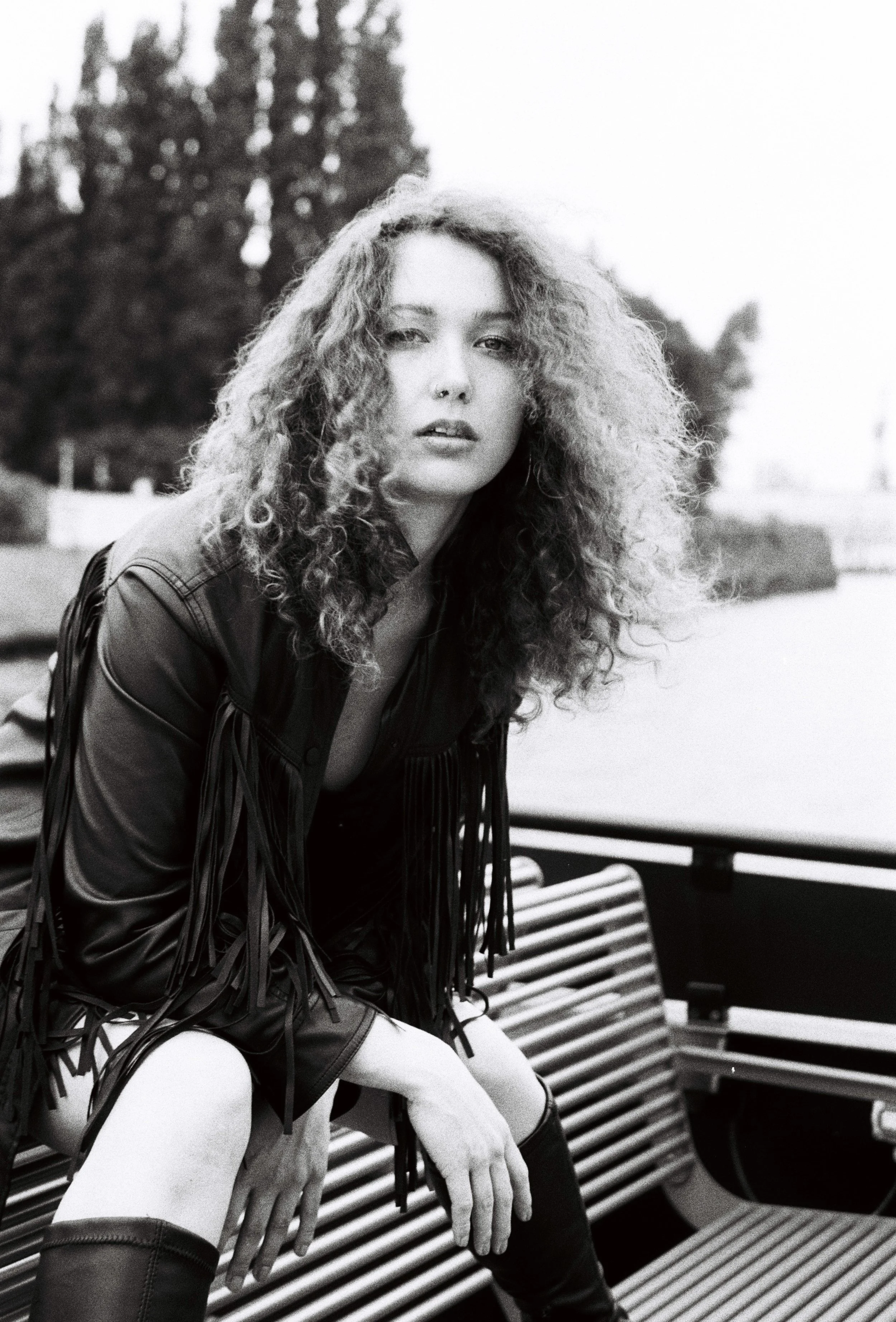 A black and white photo of a woman sitting on a park bench with a background of trees and sky. She has curly hair, is wearing thigh-high boots, a fringed jacket, and has a relaxed pose.