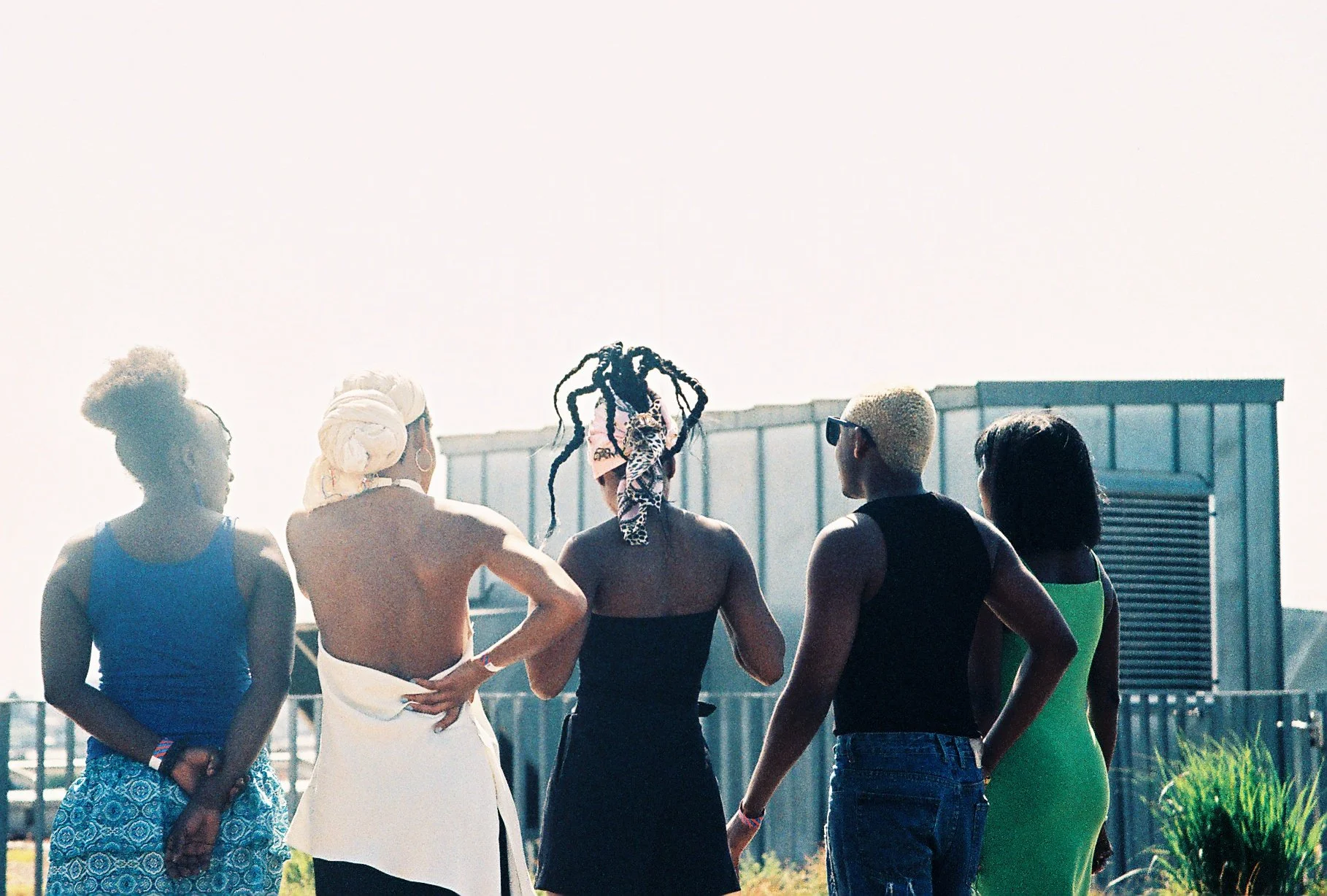A group of five women standing outdoors, facing away from the camera, with a building and sky in the background.