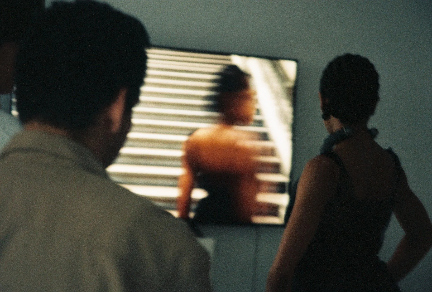 Blurred image of two people observing a woman in front of a window with horizontal blinds. The scene appears to be indoors in a dimly lit setting.