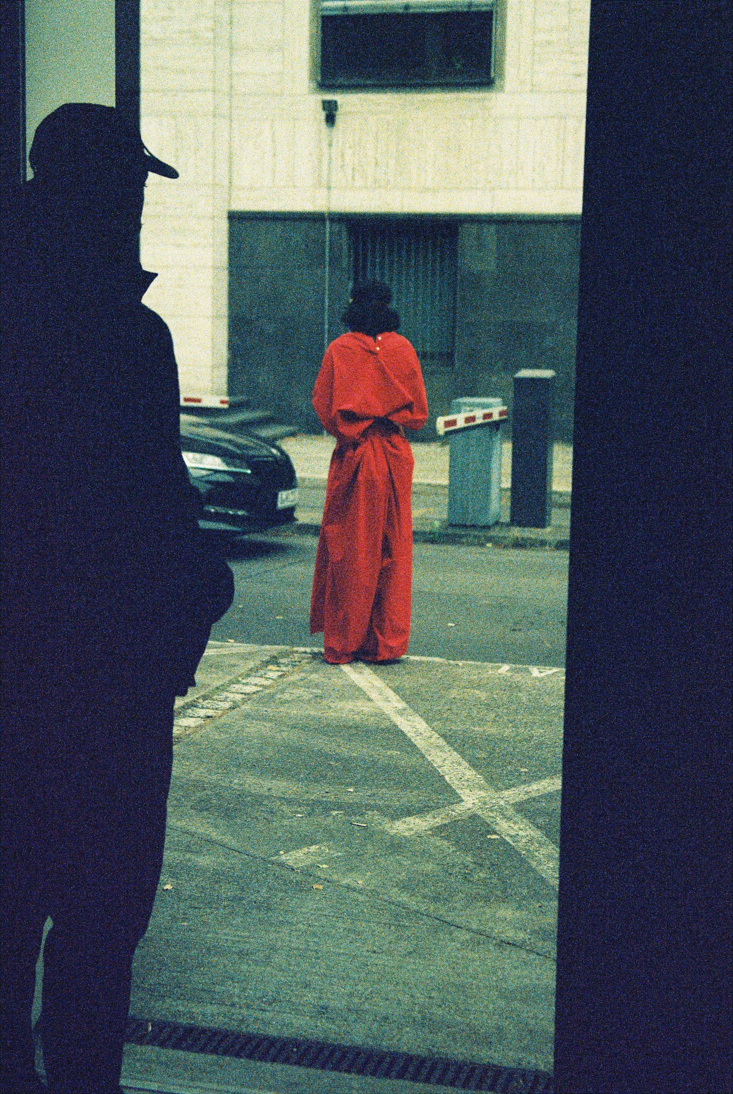 A person standing in a parking lot seen from the side, wearing a dark jacket and a cap. In the background, there is a person wearing a bright red jacket with their back turned and hands behind their back, facing a garage door. Part of a car and some 