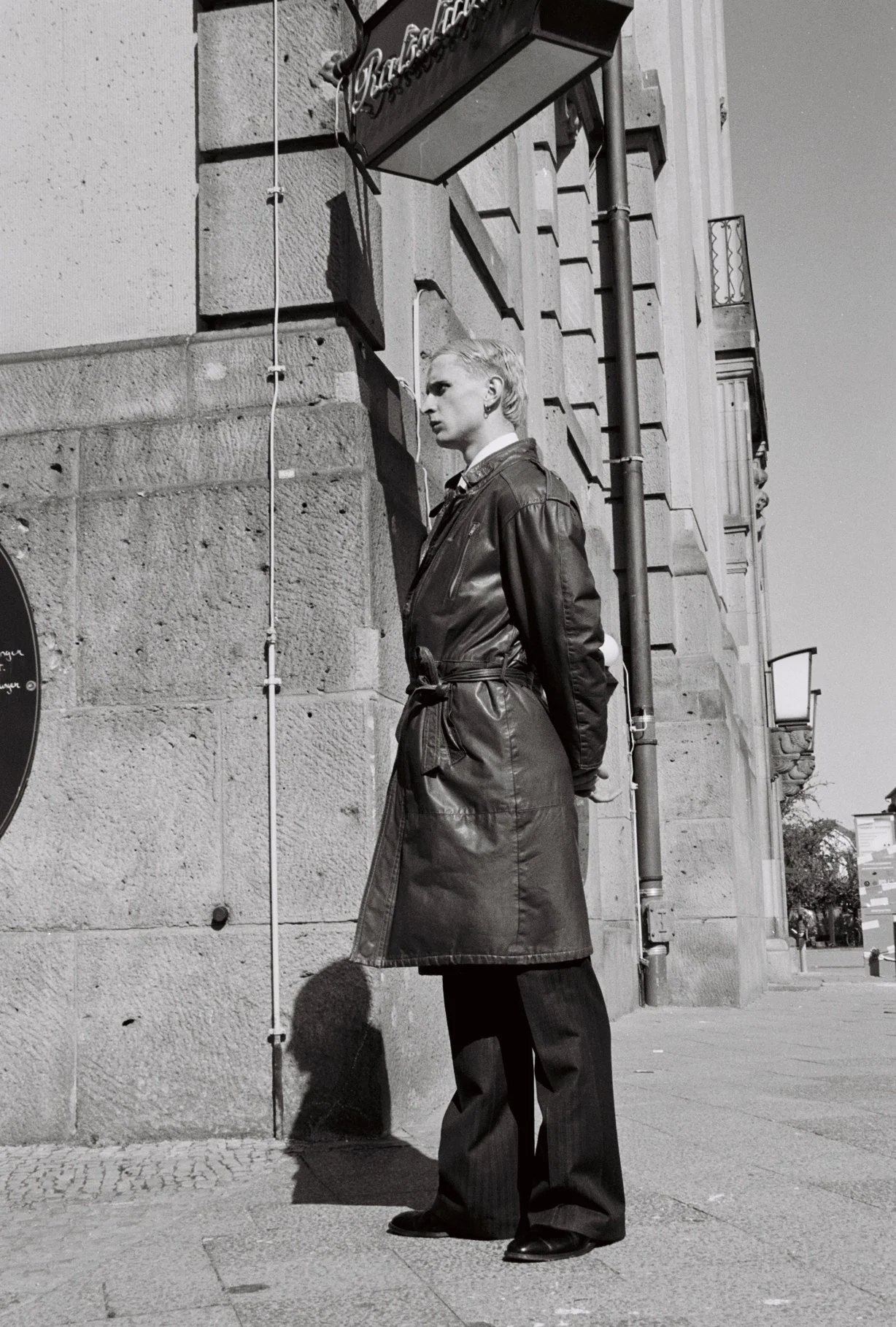 A woman stands on a city sidewalk against a stone building wall, oriented sideways, gazing into the distance. She wears a long leather coat, dark pants, and heels, with her hair styled back.