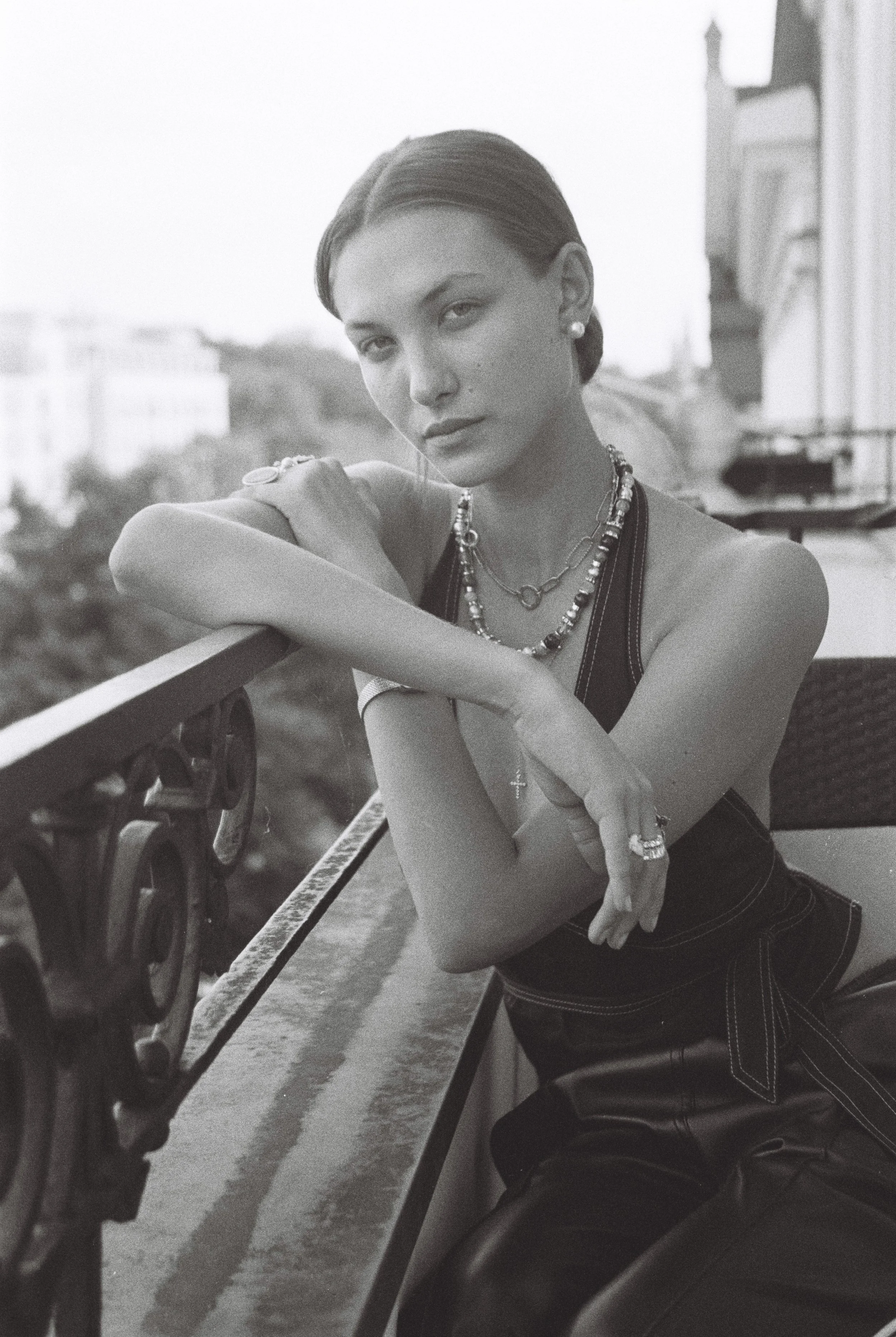 A young woman with dark hair in a side bun, wearing a sleeveless top, beaded necklaces, and earrings, leaning on a city balcony railing, looking at the camera with a serious expression, in black and white.