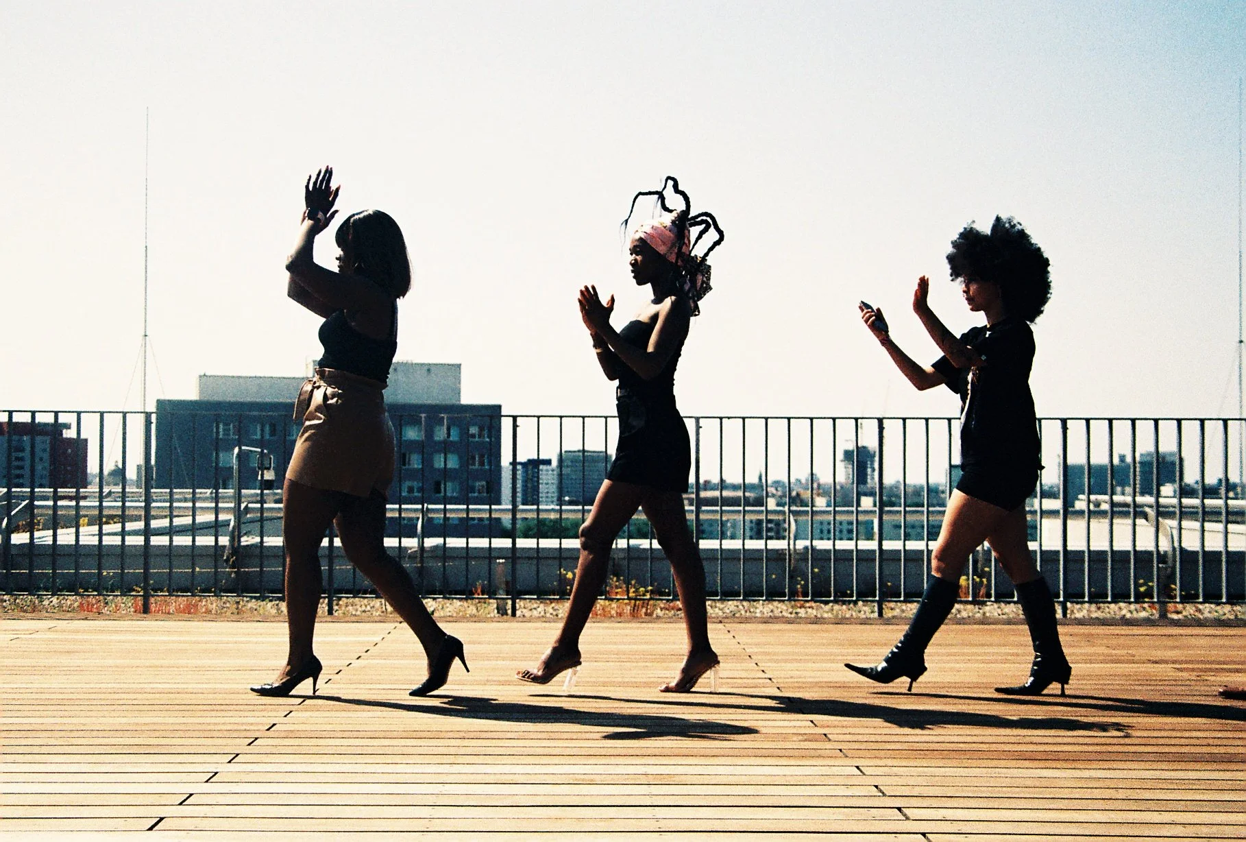 Three women walking on a rooftop deck, dressed in stylish outfits and high heels, with an urban cityscape in the background.