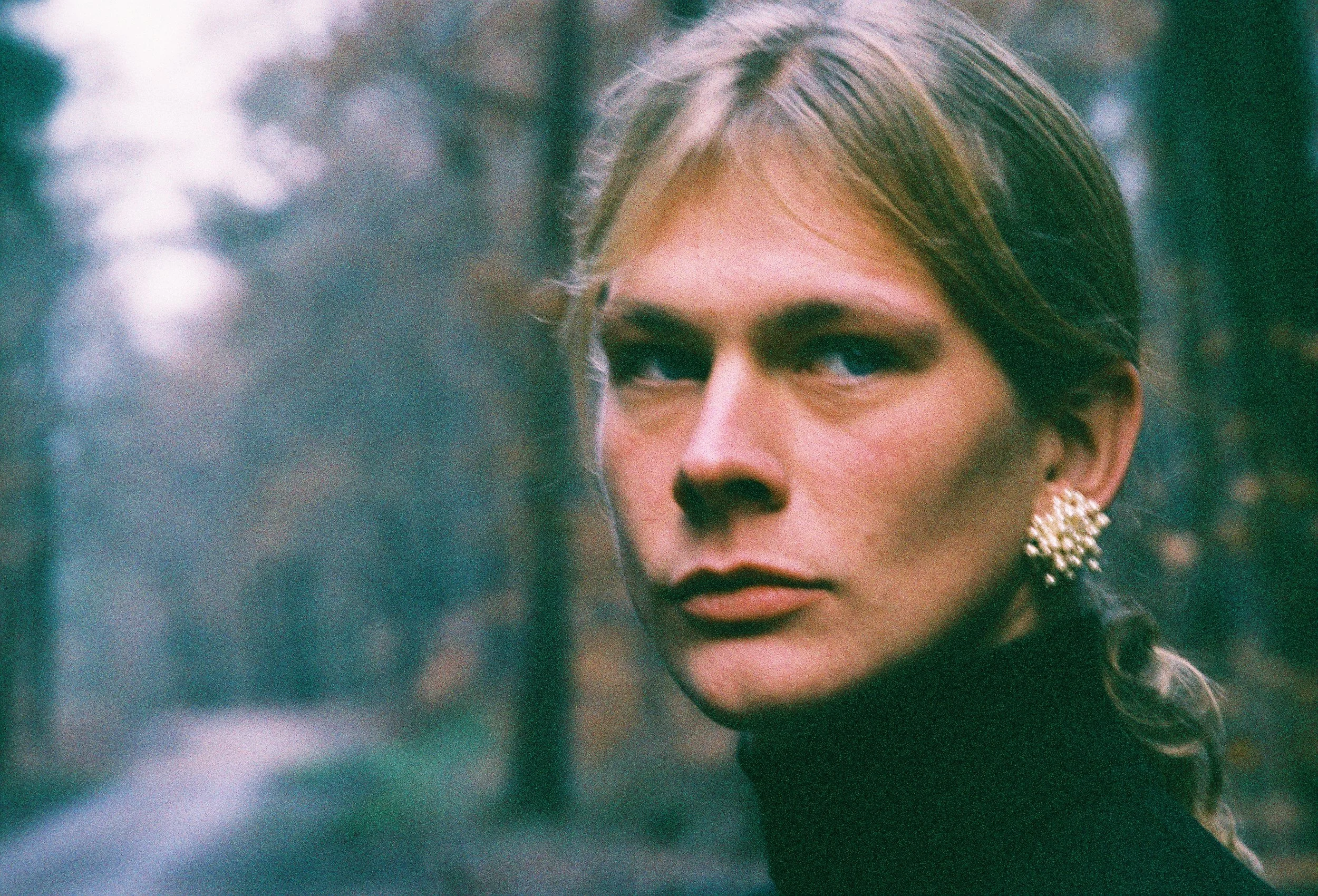 Close-up of a woman's face, showing her eye, part of her nose, and earrings, with a blurred outdoor background.