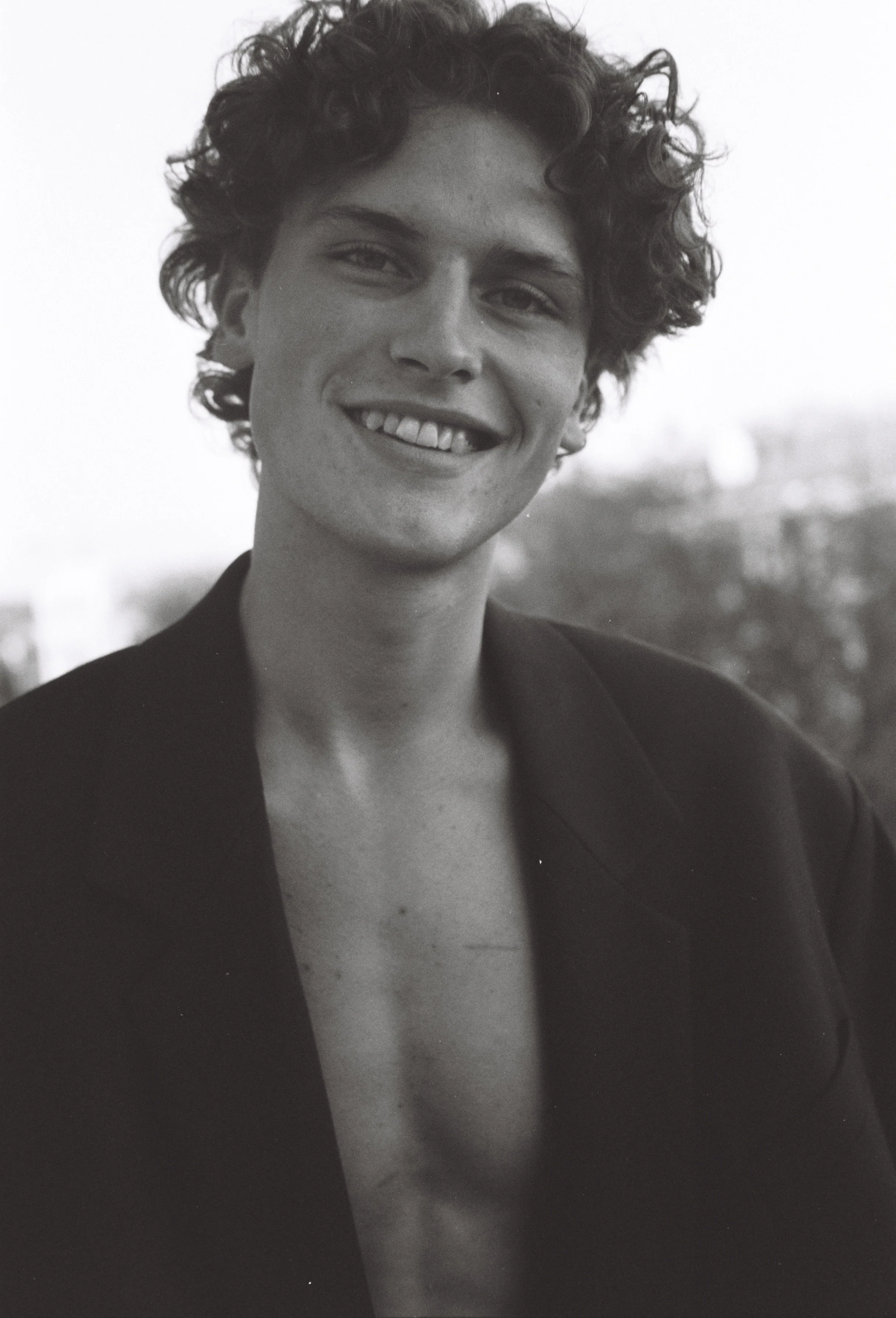 A black-and-white portrait photograph of a smiling woman with short, curly hair, wearing a dark blazer, outdoors with a blurred cityscape background.