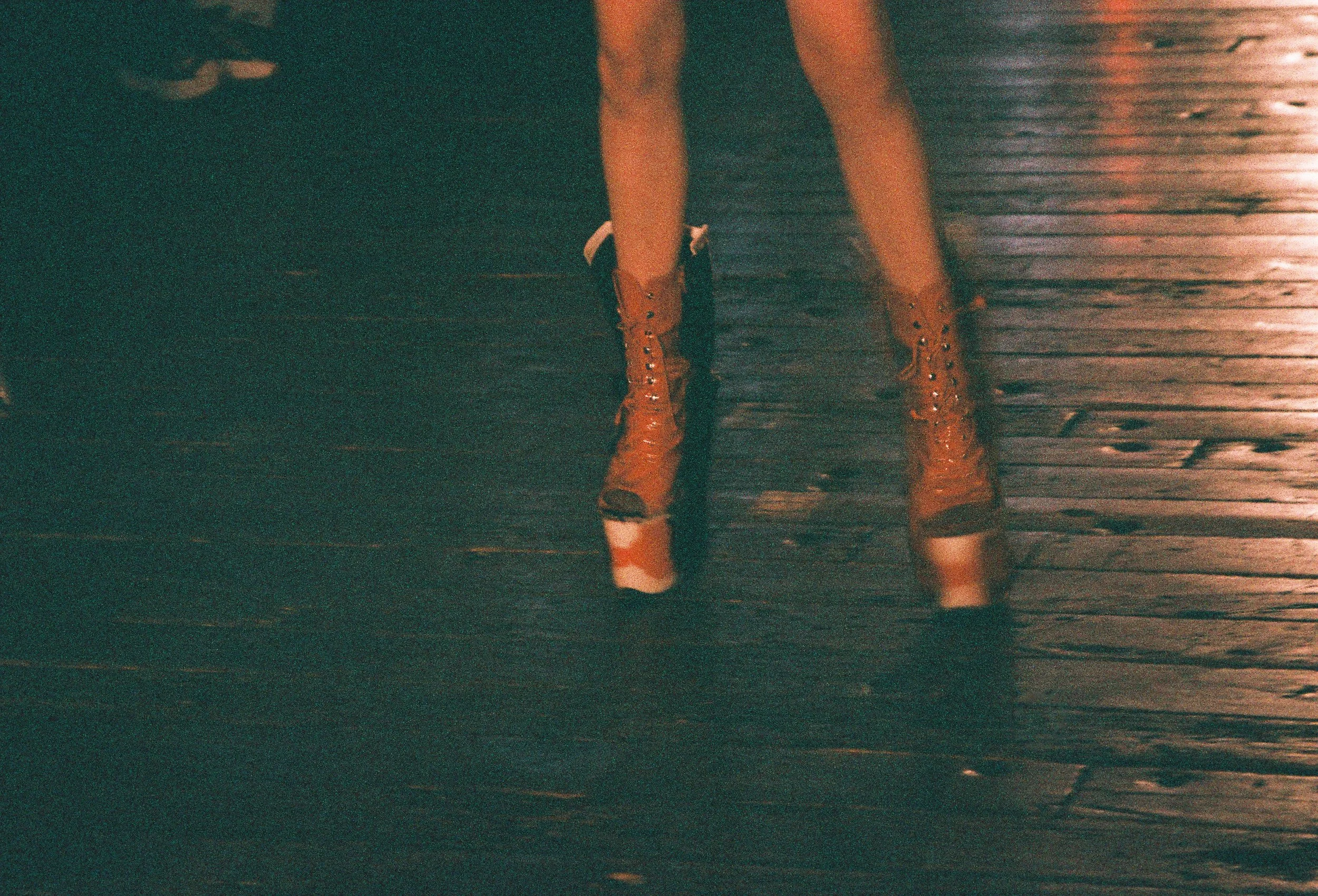Lower half of a person standing on a wet wooden deck at night, wearing tan work boots and shorts, with water splashing around their feet.