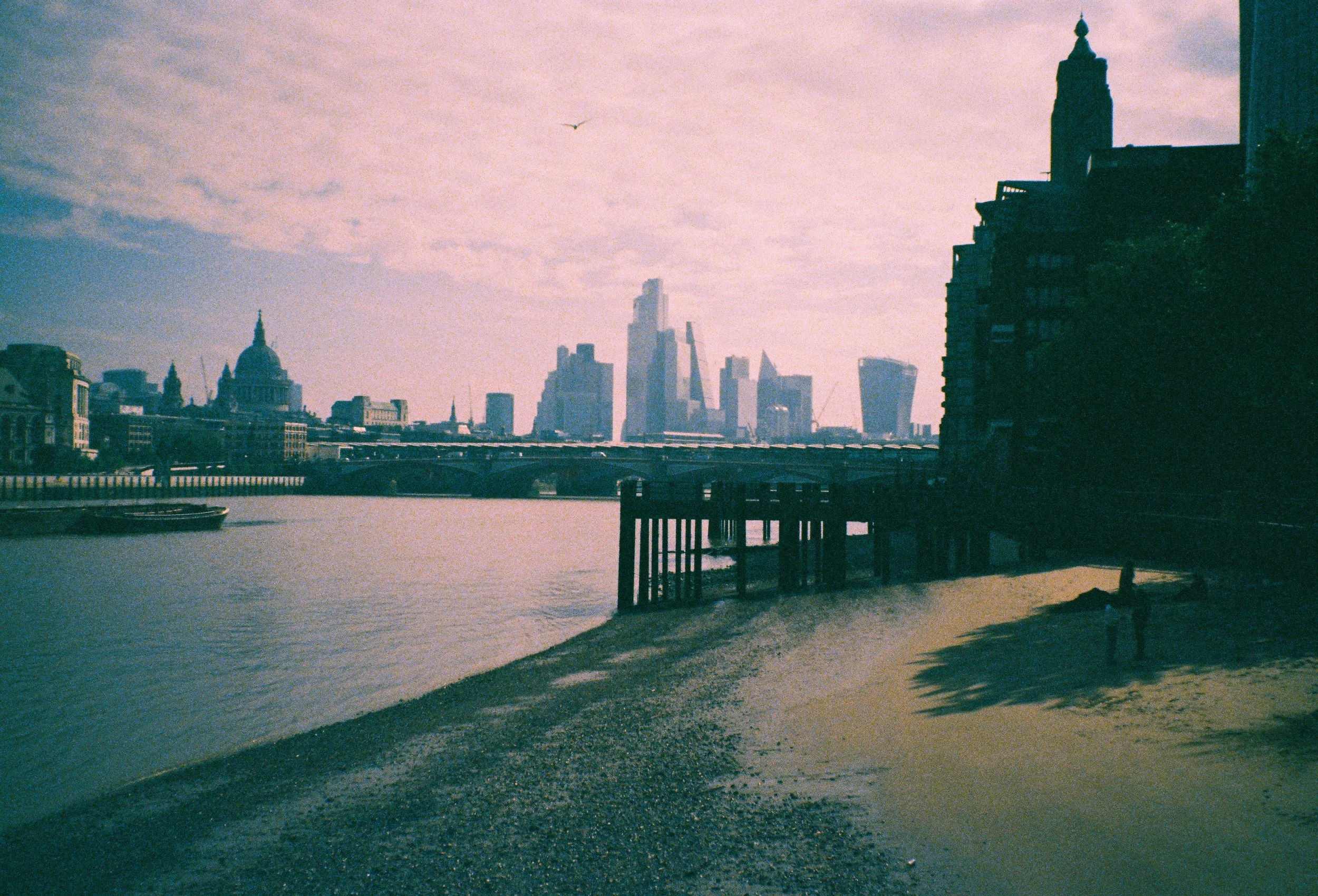 A city skyline with tall buildings and a river in the foreground, during sunset or sunrise, with a cloudy sky.