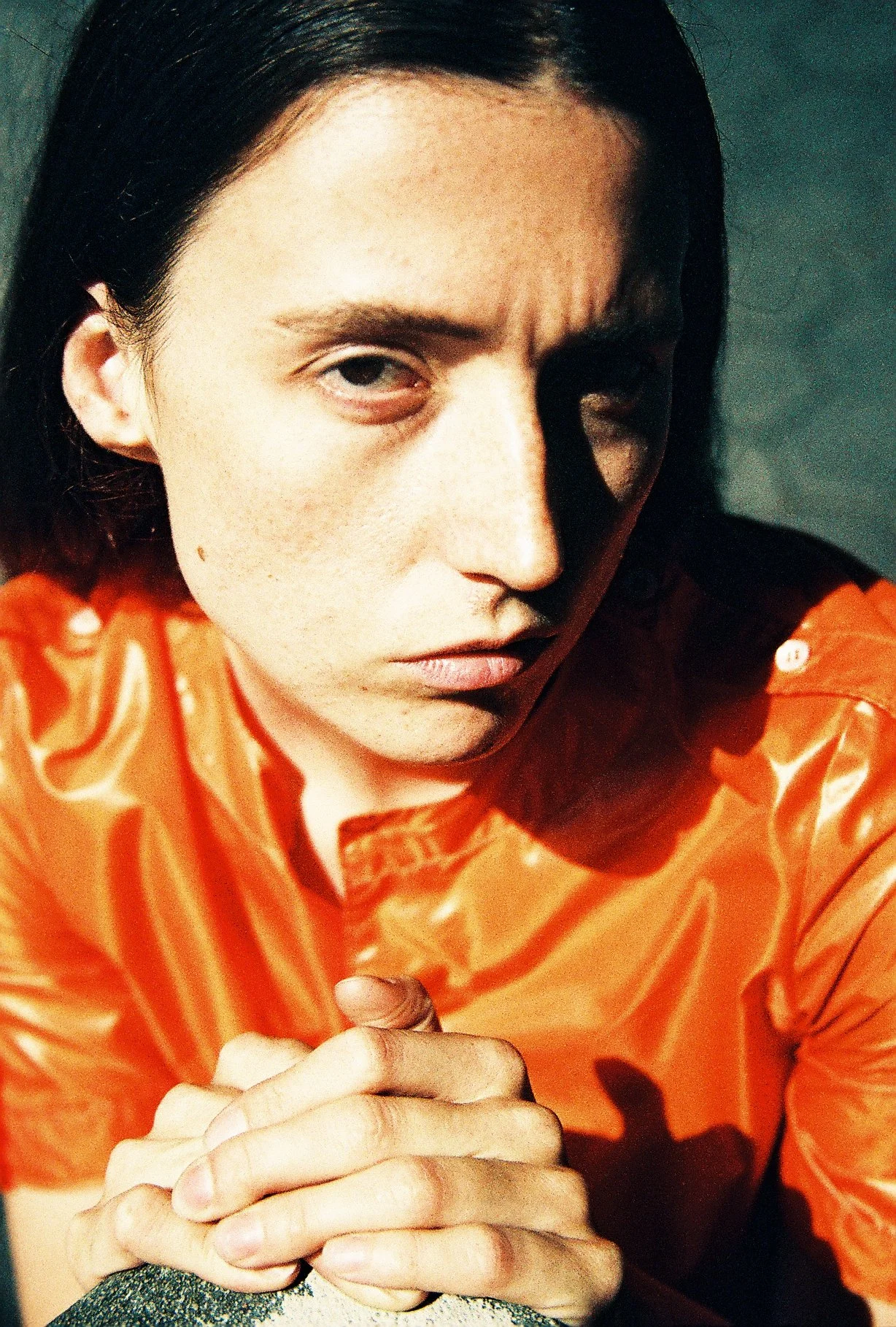 A person with dark hair and light skin is wearing a bright orange satin shirt and looking intently at the camera. They have a serious expression and are clasping their hands together close to their face.