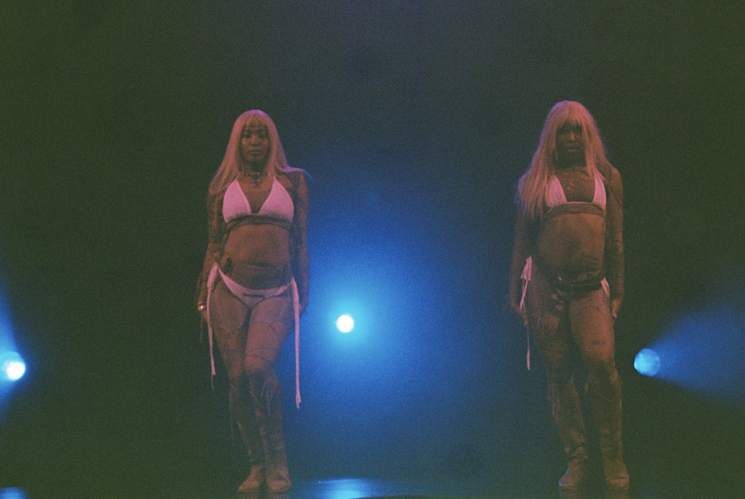Two women with long blonde hair standing side by side on a dark stage, illuminated by blue and purple lighting, wearing matching athletic outfits.