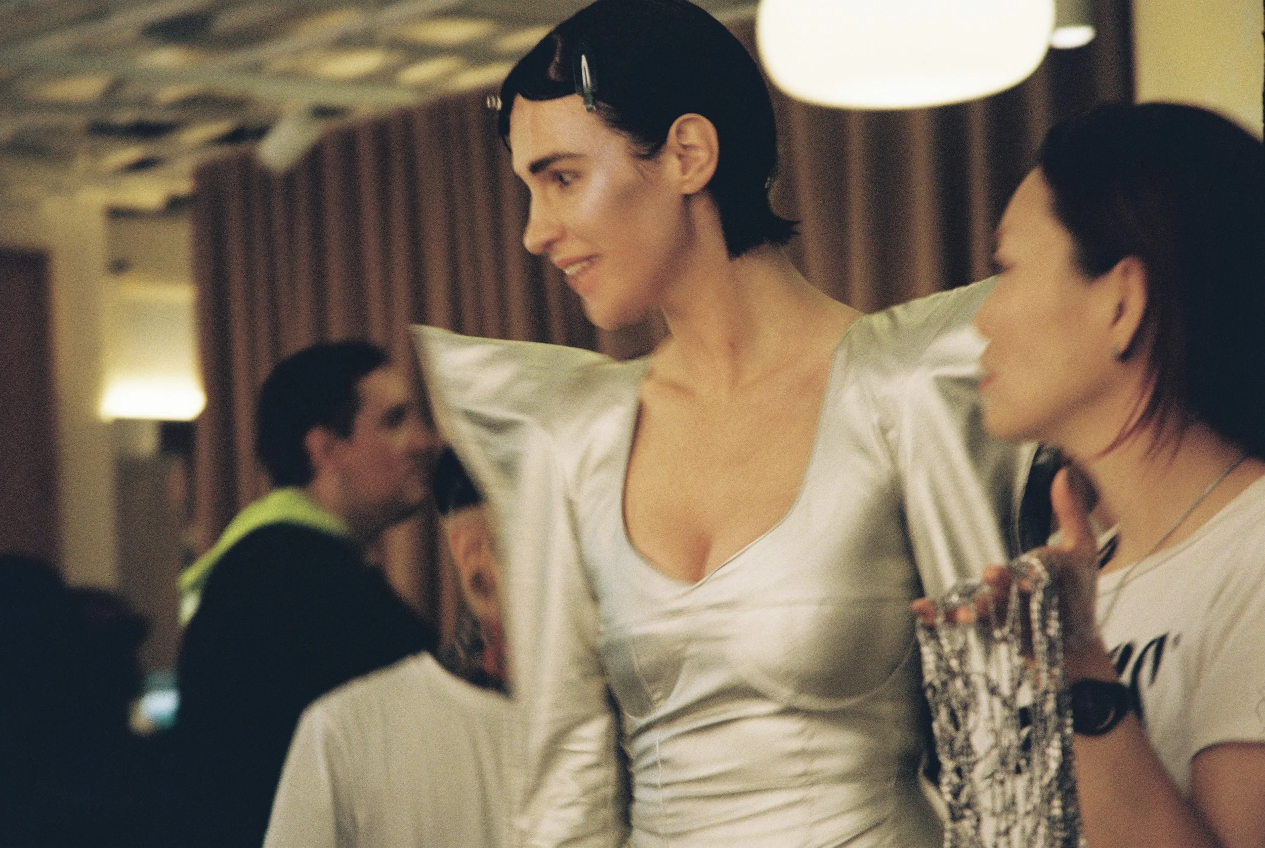 A woman with short dark hair, wearing a white shirt, smiling and looking down at a woman with long dark hair and glasses, who is wearing a patterned top, in an indoor setting with warm lighting.