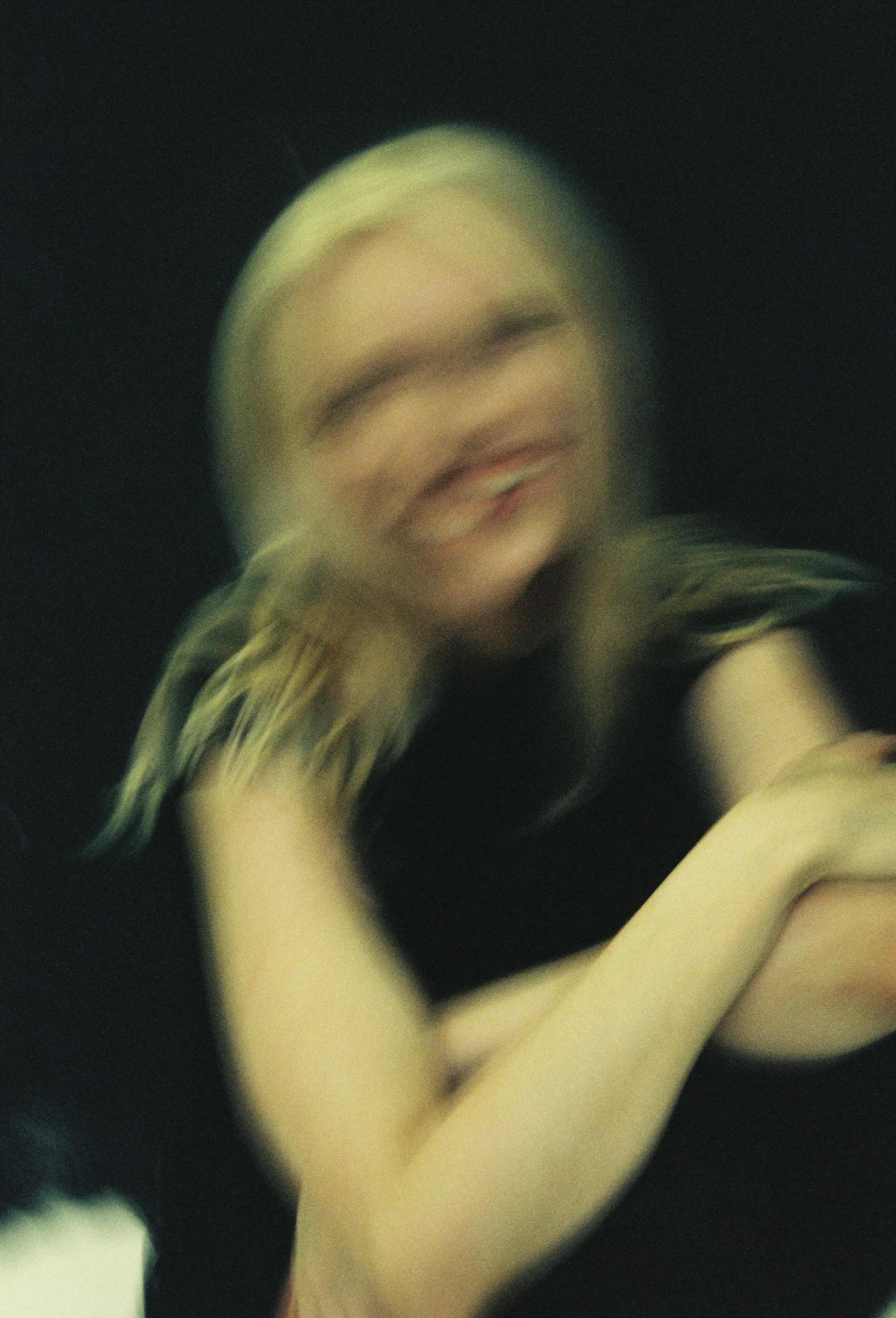 A blurry close-up photo of a woman with blonde hair, smiling with her eyes closed, against a dark background.