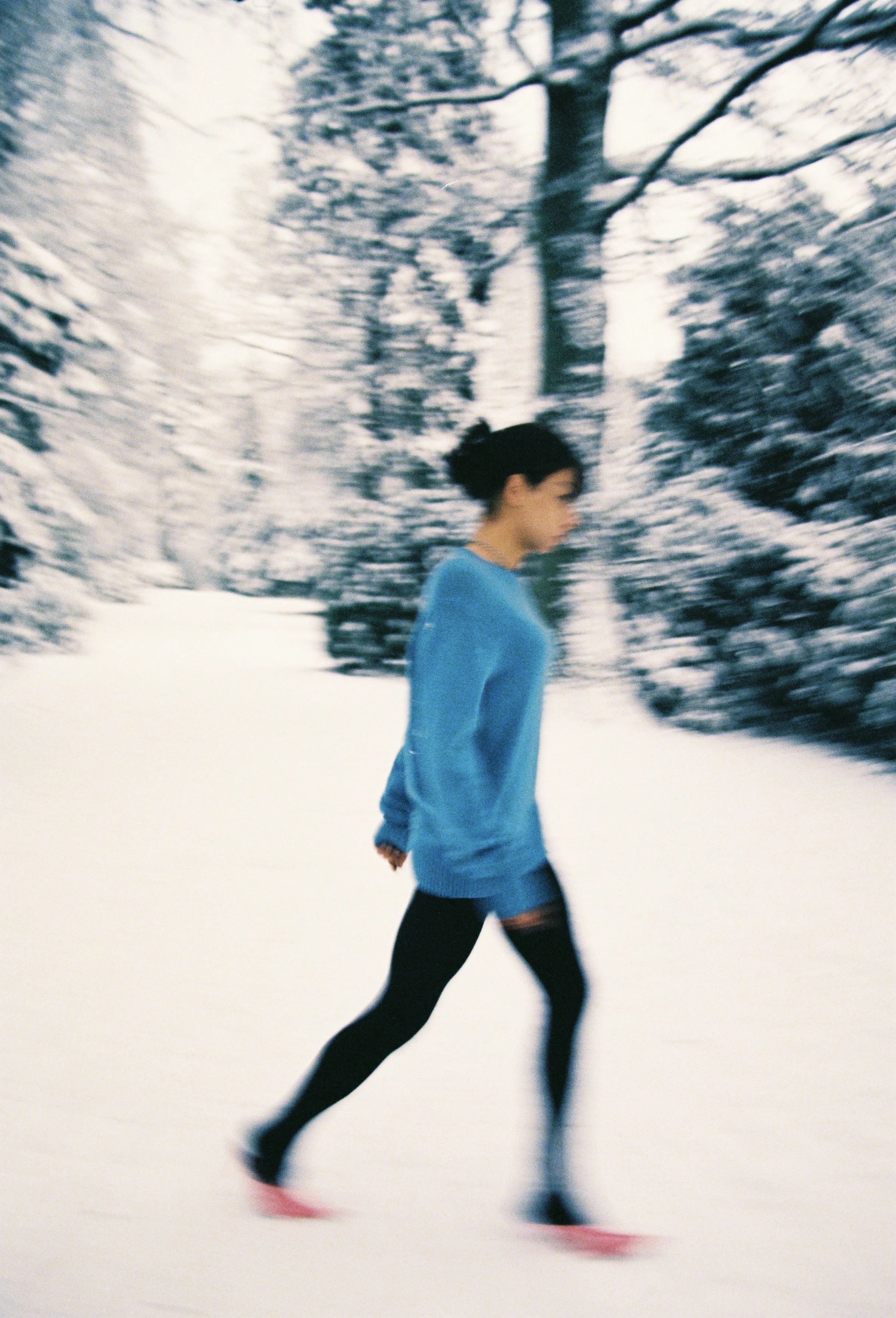 A woman in a blue jacket and black leggings is running on snow-covered ground in a winter landscape with snow-covered trees.