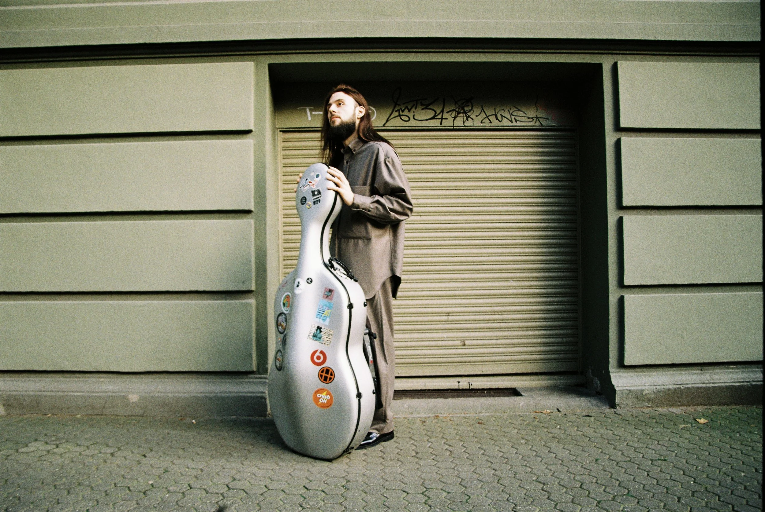 A man with long hair and a beard stands on a sidewalk holding a large guitar case covered in stickers. He is dressed in a long, brown coat and dark shoes, positioned against a green wall with a closed, graffiti-covered shutter.