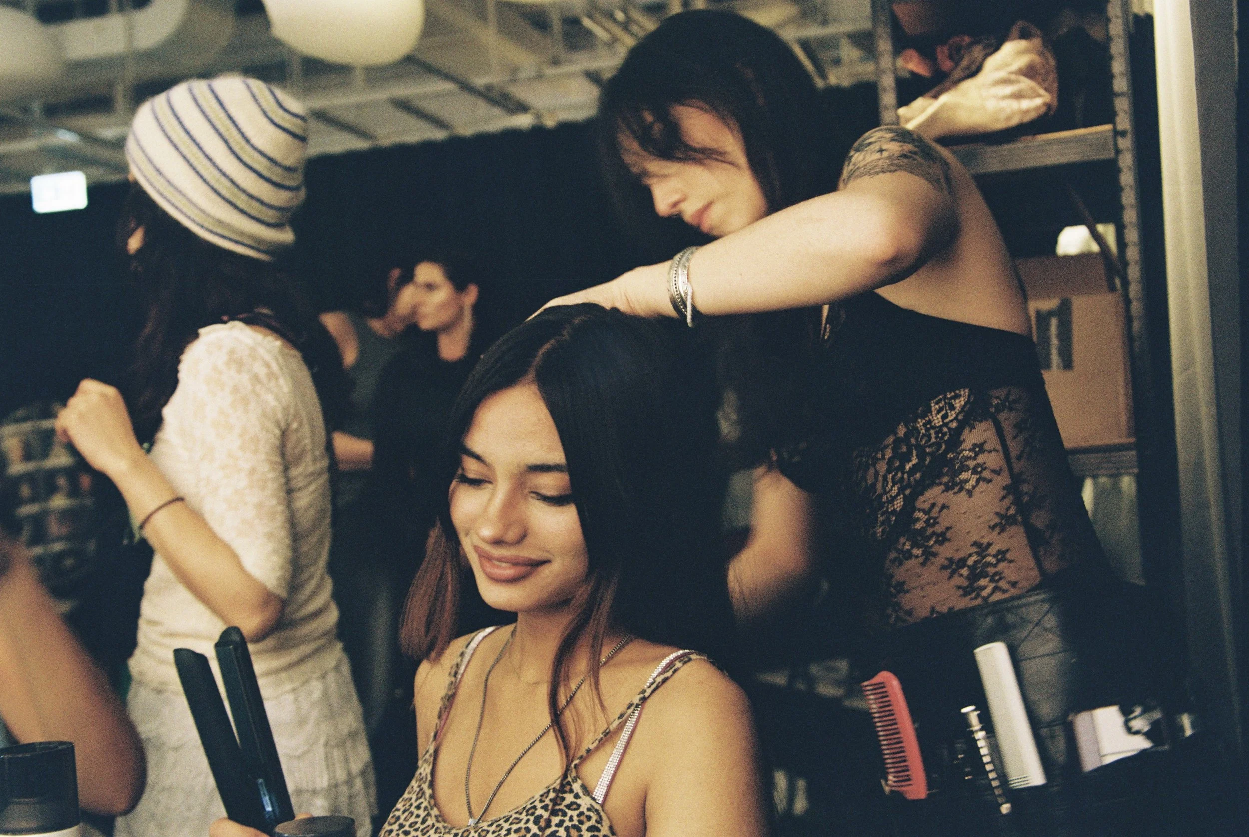 A woman with long brown hair and a leopard print top smiling with her eyes closed while another woman with black hair and a tattooed arm styled in dark clothing styles her hair at a salon, with other people and salon equipment visible in the backgrou