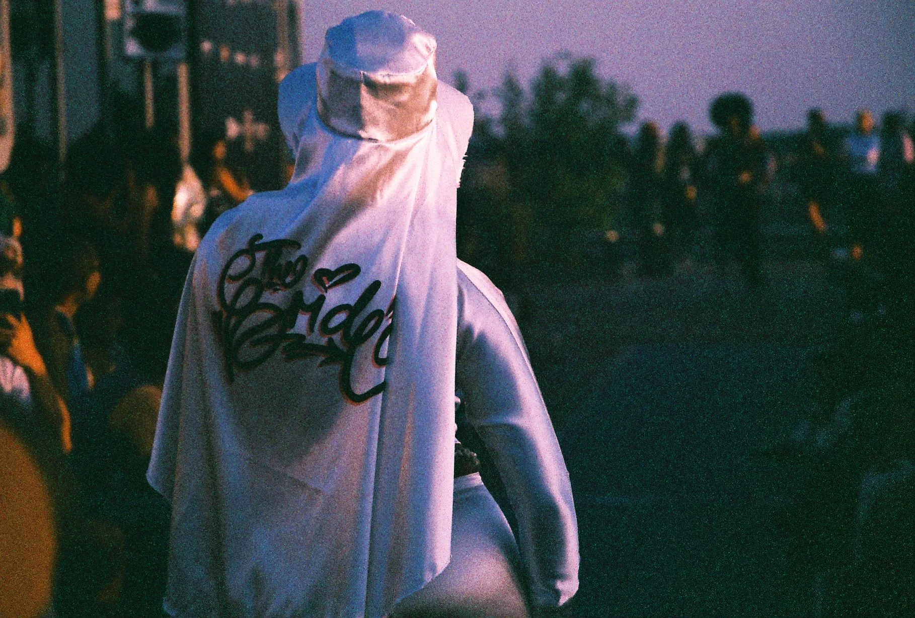 A person wearing a wide-brimmed hat and a white, loose-fitting shirt with graffiti-like writing and a heart on the back, standing outdoors during dusk with a crowd of people and trees in the background.
